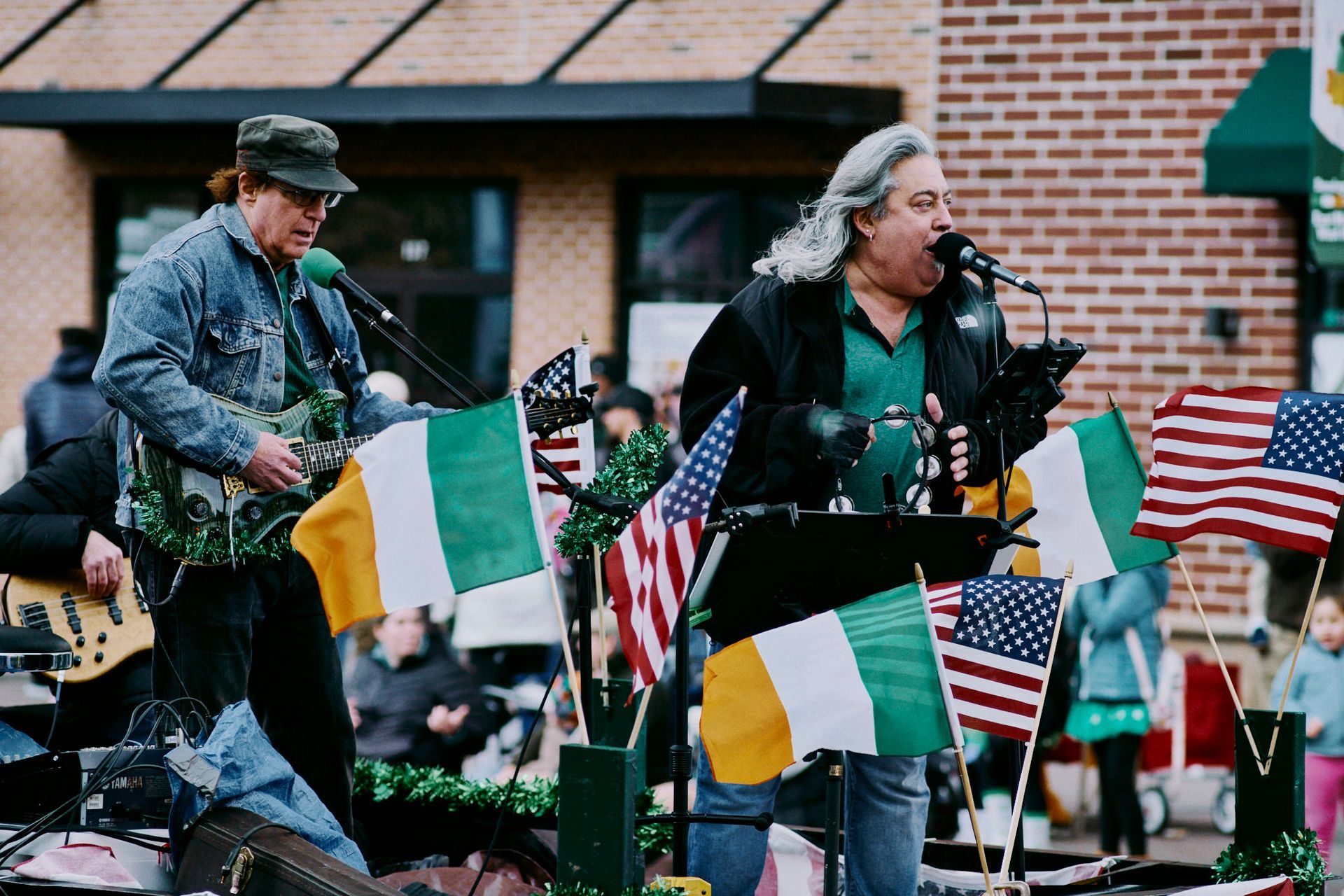 Two men playing music, singing into microphones outdoors, surrounded by Irish and American flags.