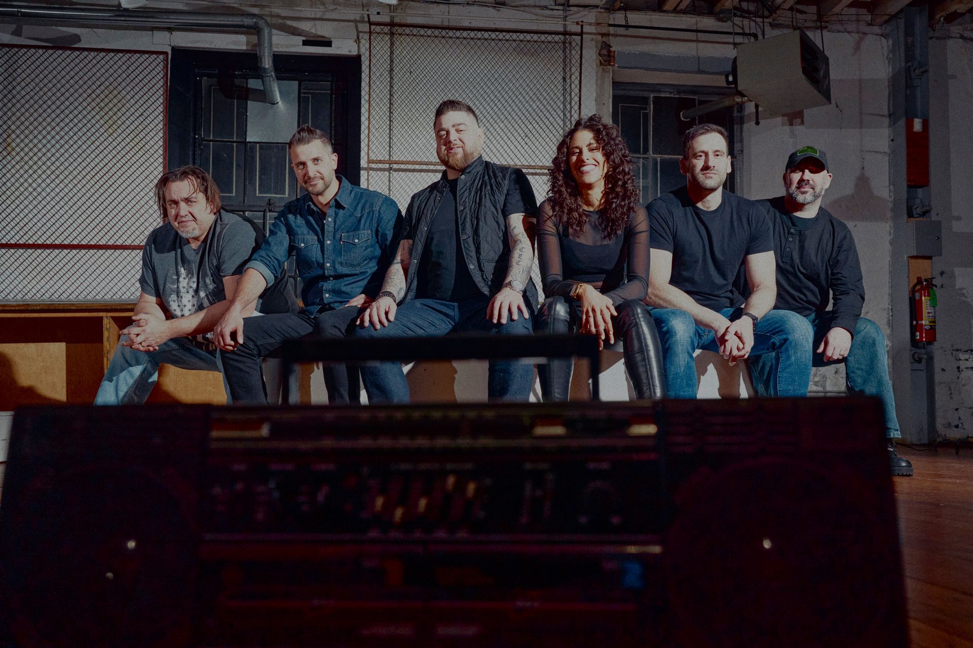 Band posing on a bench in a studio. Five people: two men, a woman, and two men. Dark, industrial setting.