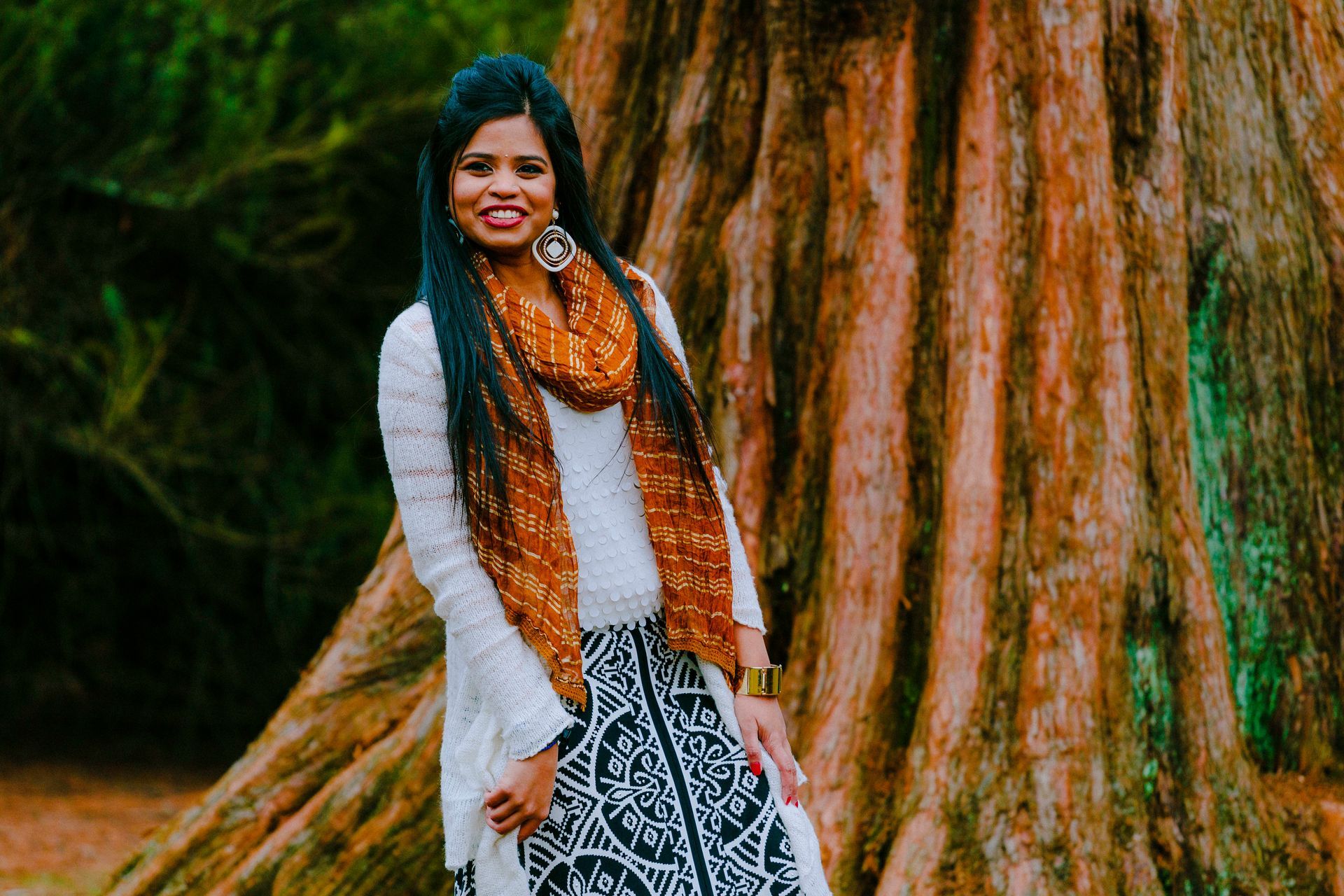 Woman smiling next to a large tree; wearing a white sweater, patterned skirt, and orange scarf.