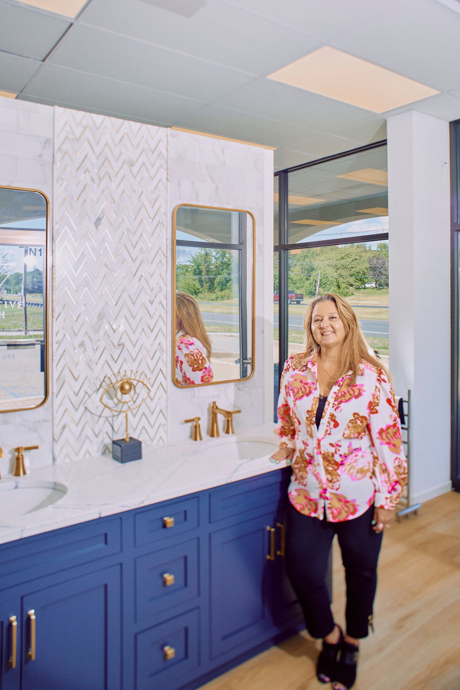 Woman in floral shirt stands near a blue bathroom vanity with gold accents and a white backsplash.