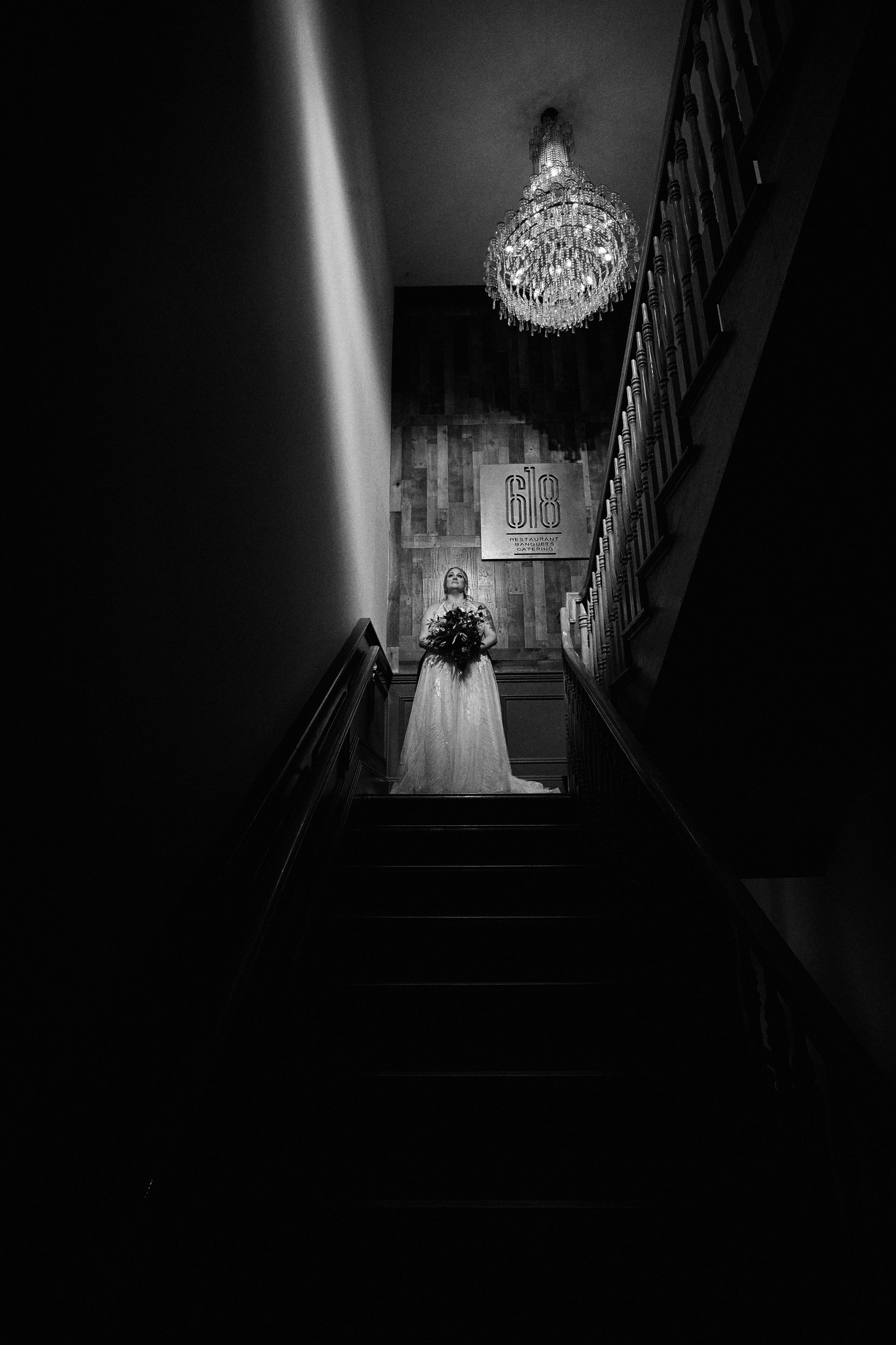 Bride in a white dress stands at the bottom of a dark staircase under a chandelier, black and white.