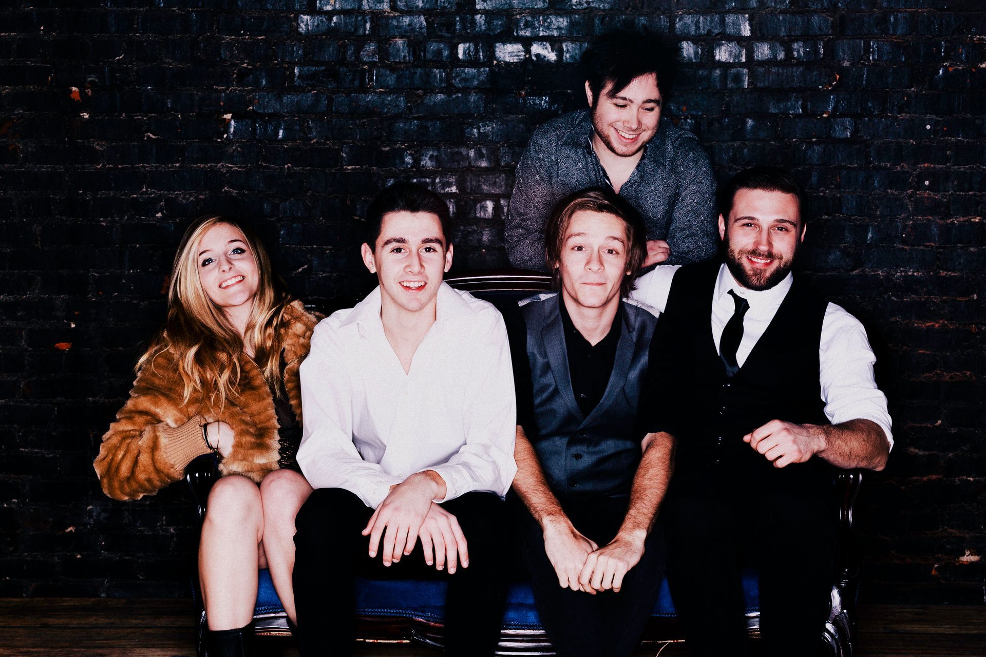 Band of five smiling, posing in front of a brick wall.
