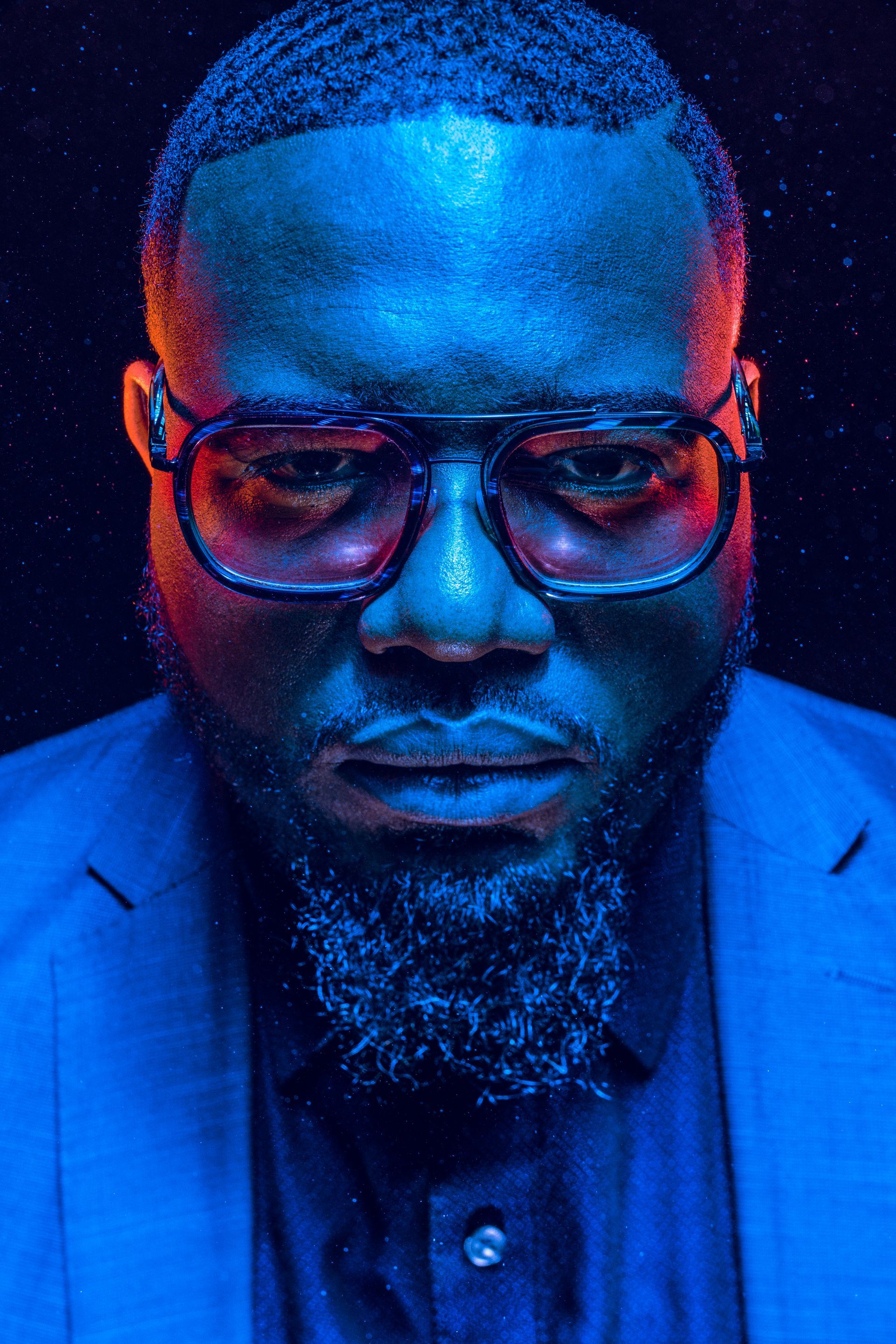 Man in blue suit and glasses, lit with red and blue light, water droplets.