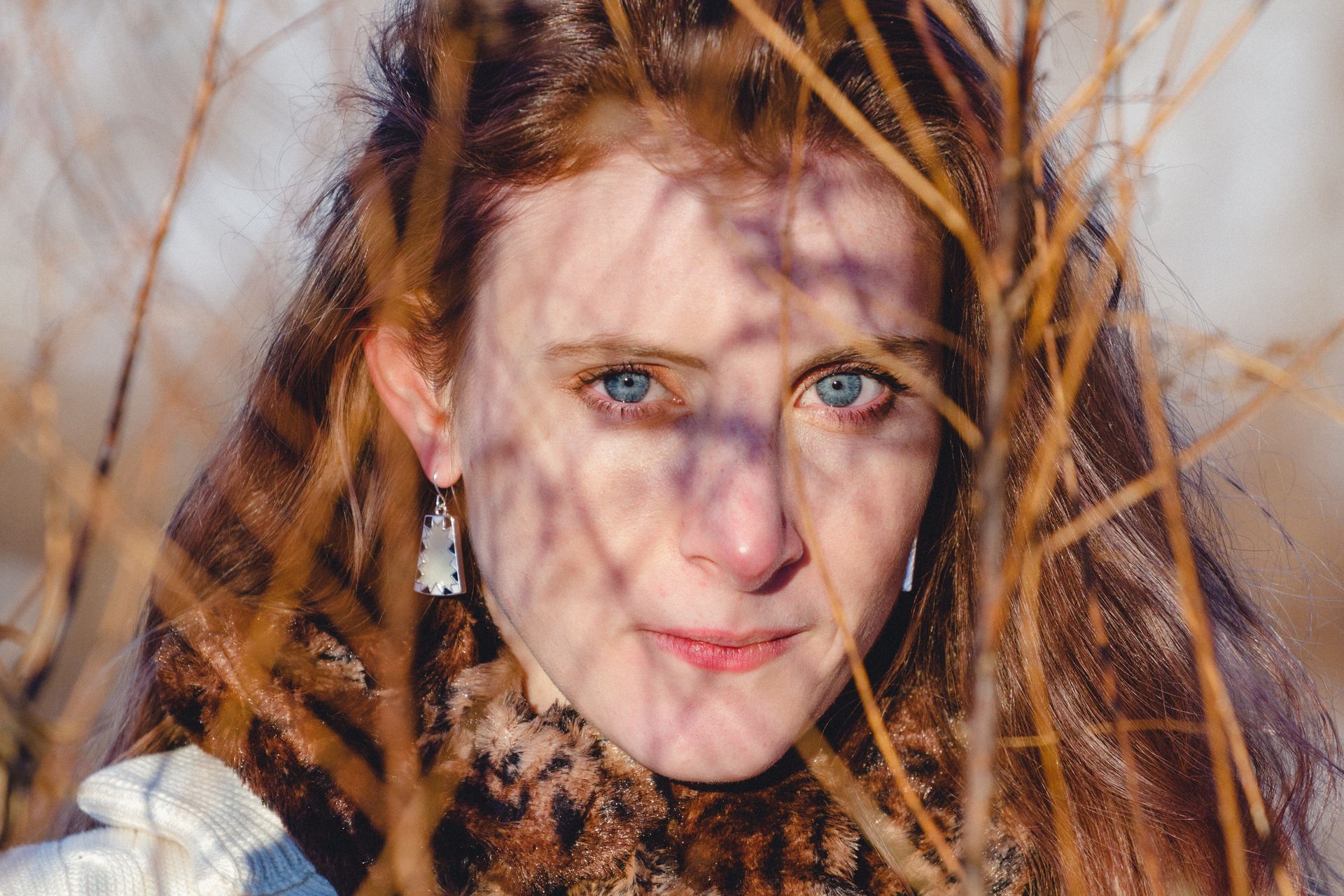 Woman with red hair, blue eyes, and light skin peeking through brown branches in a natural setting.