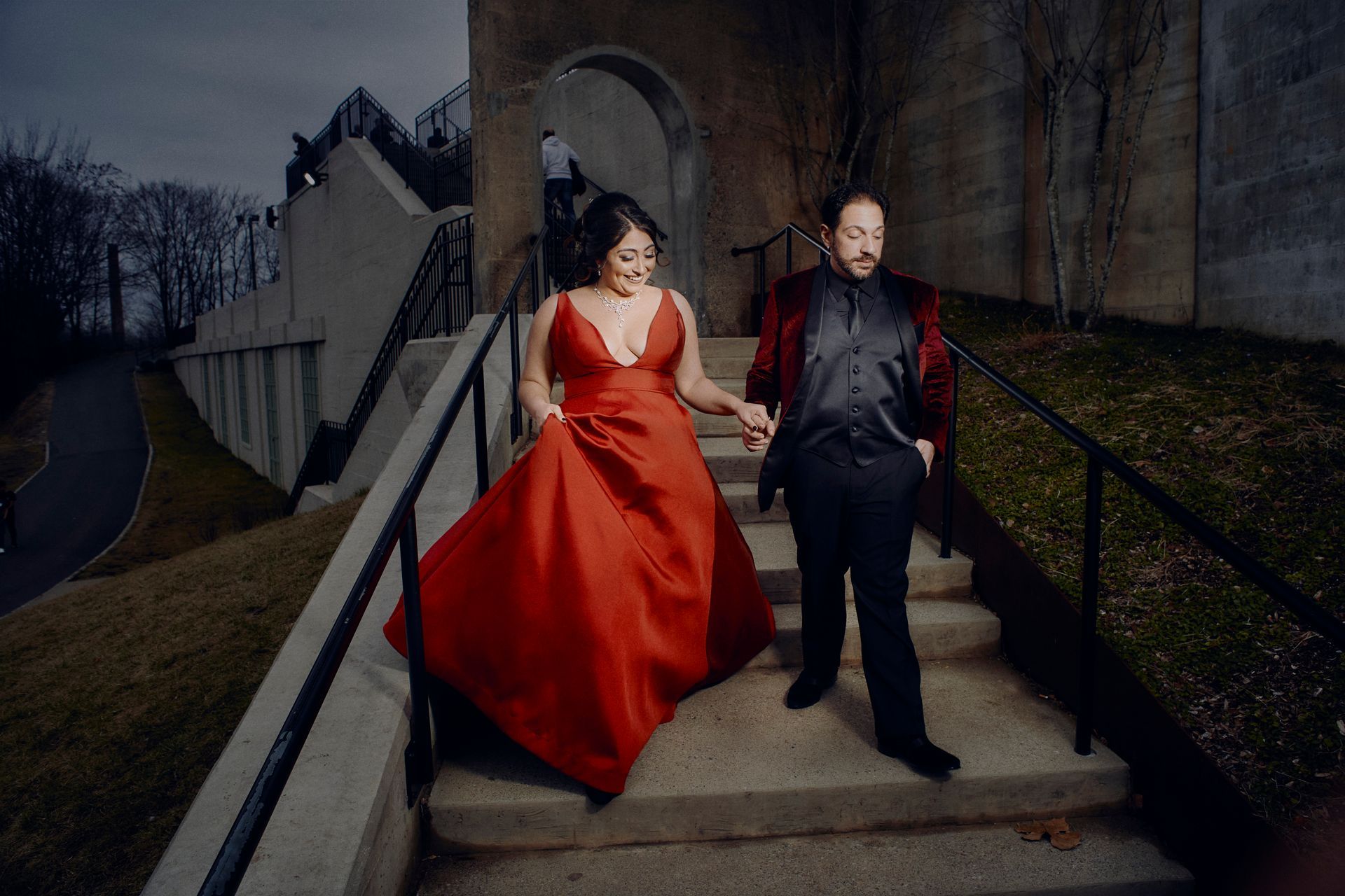 Couple descends concrete stairs; woman in red gown, man in black suit, dark background.