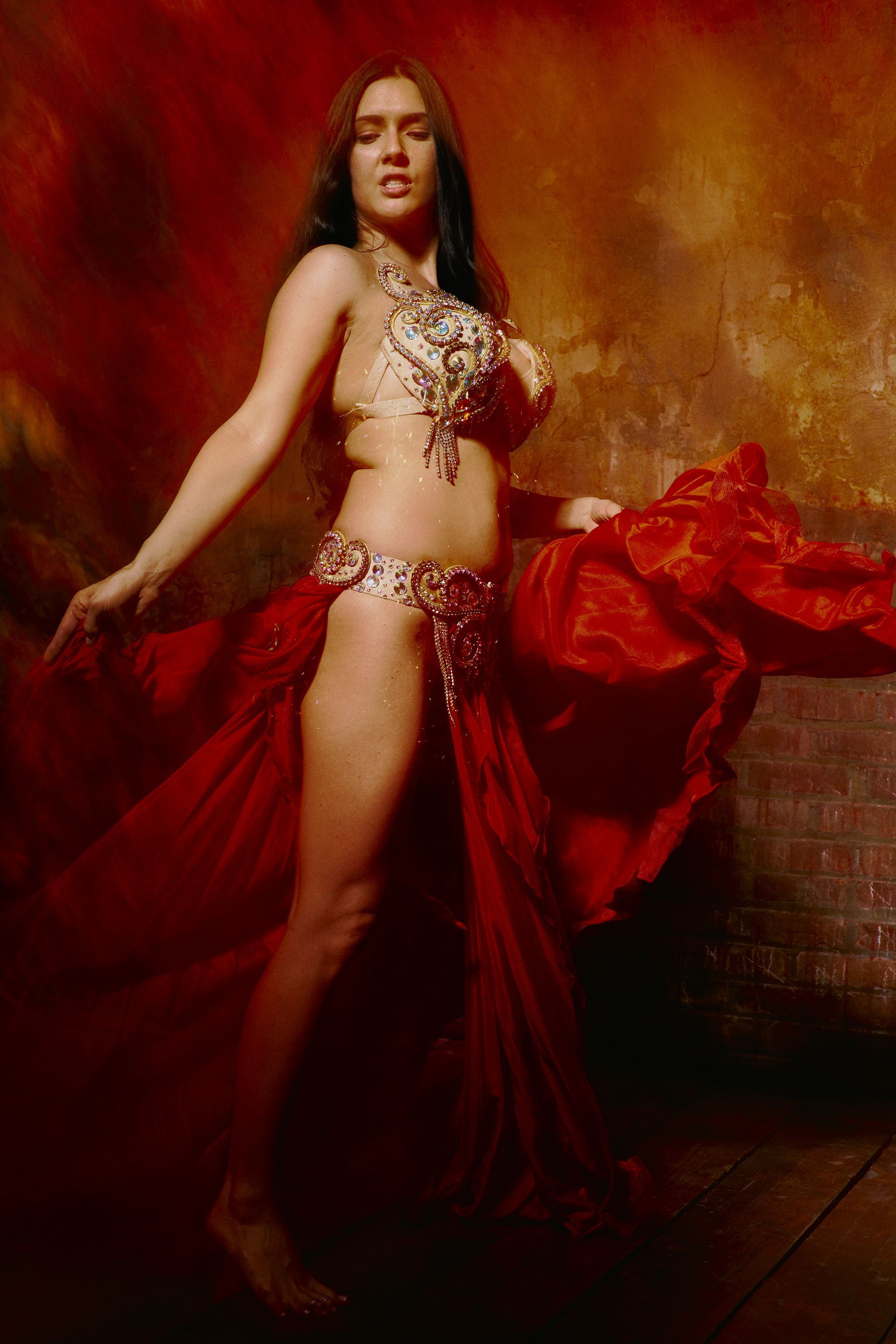 Woman in red belly dancing costume, arms outstretched, posing in front of a warm backdrop.