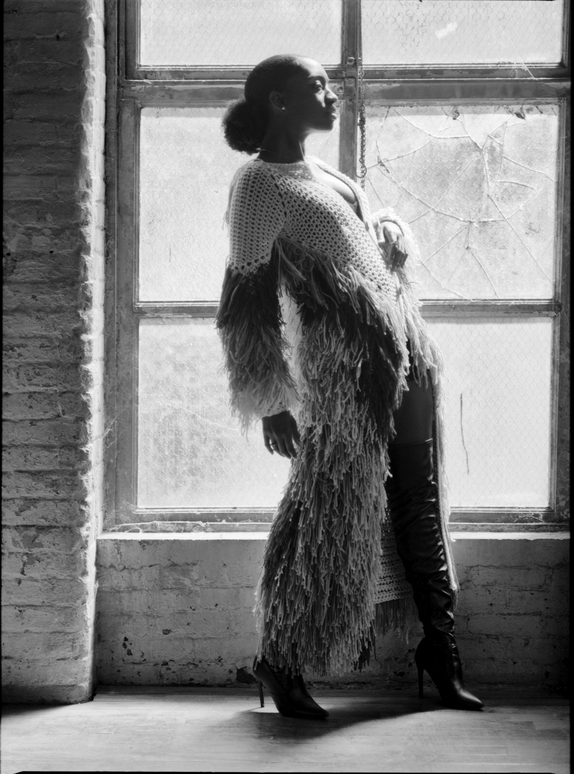 Woman in shaggy coat and boots, standing by a window, looking out. Black and white.