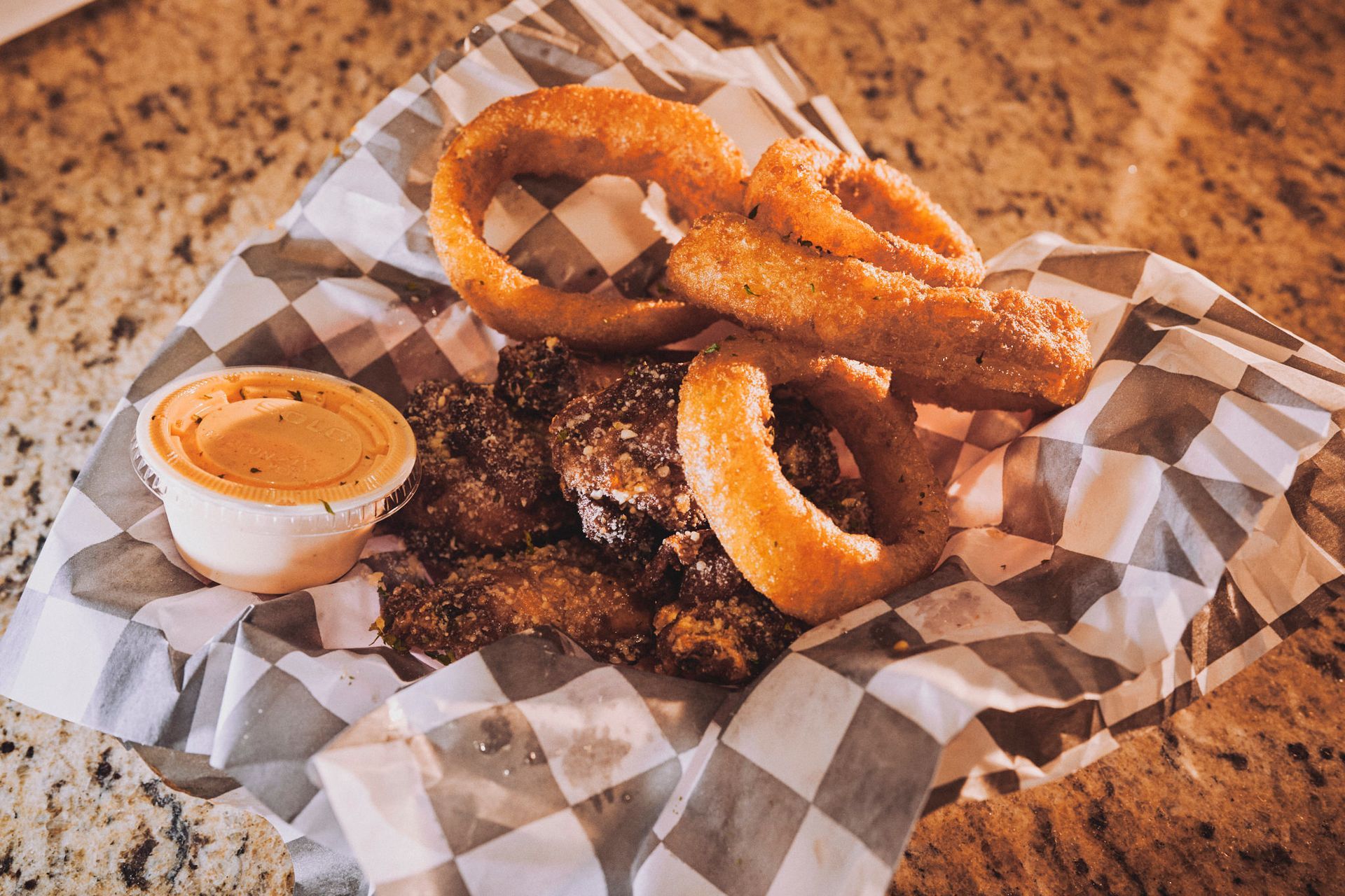 A basket of onion rings and chicken wings on a checkered paper on a table.