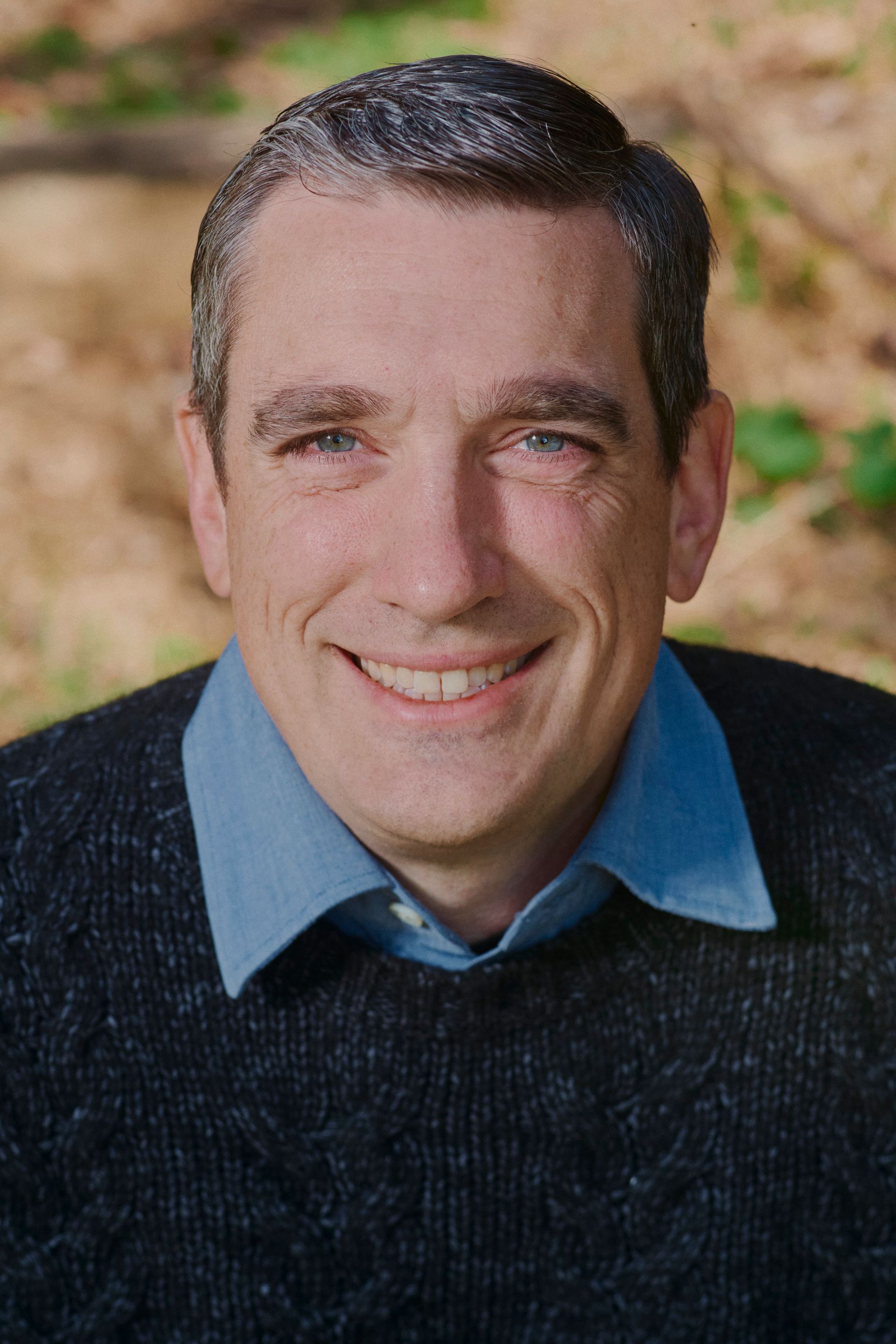 Man with graying hair smiles at the camera. Wearing a blue shirt under a dark sweater. Outdoors, blurred background.