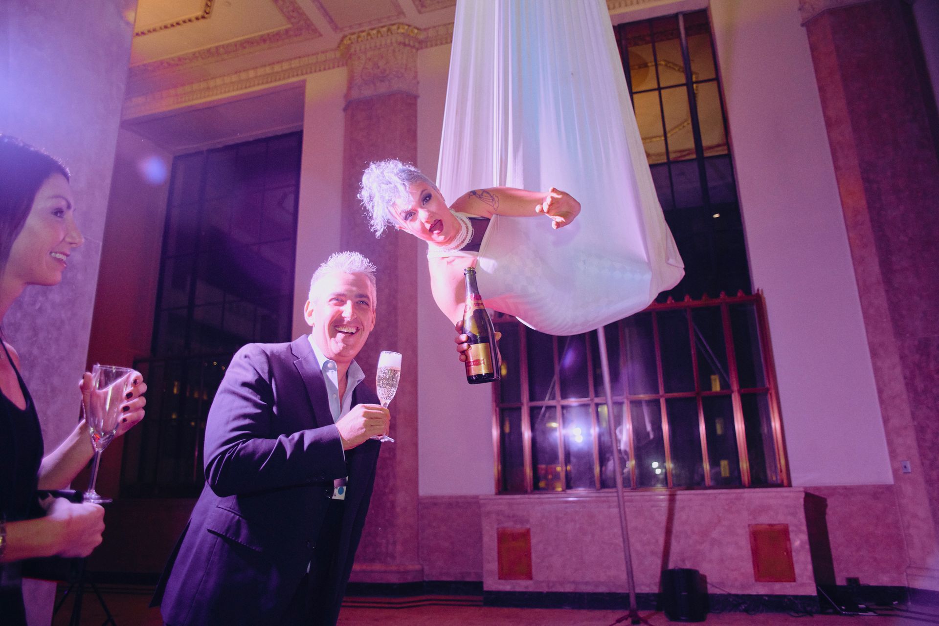 Man with champagne smiles at performer suspended in a white fabric, holding champagne bottle at an event.