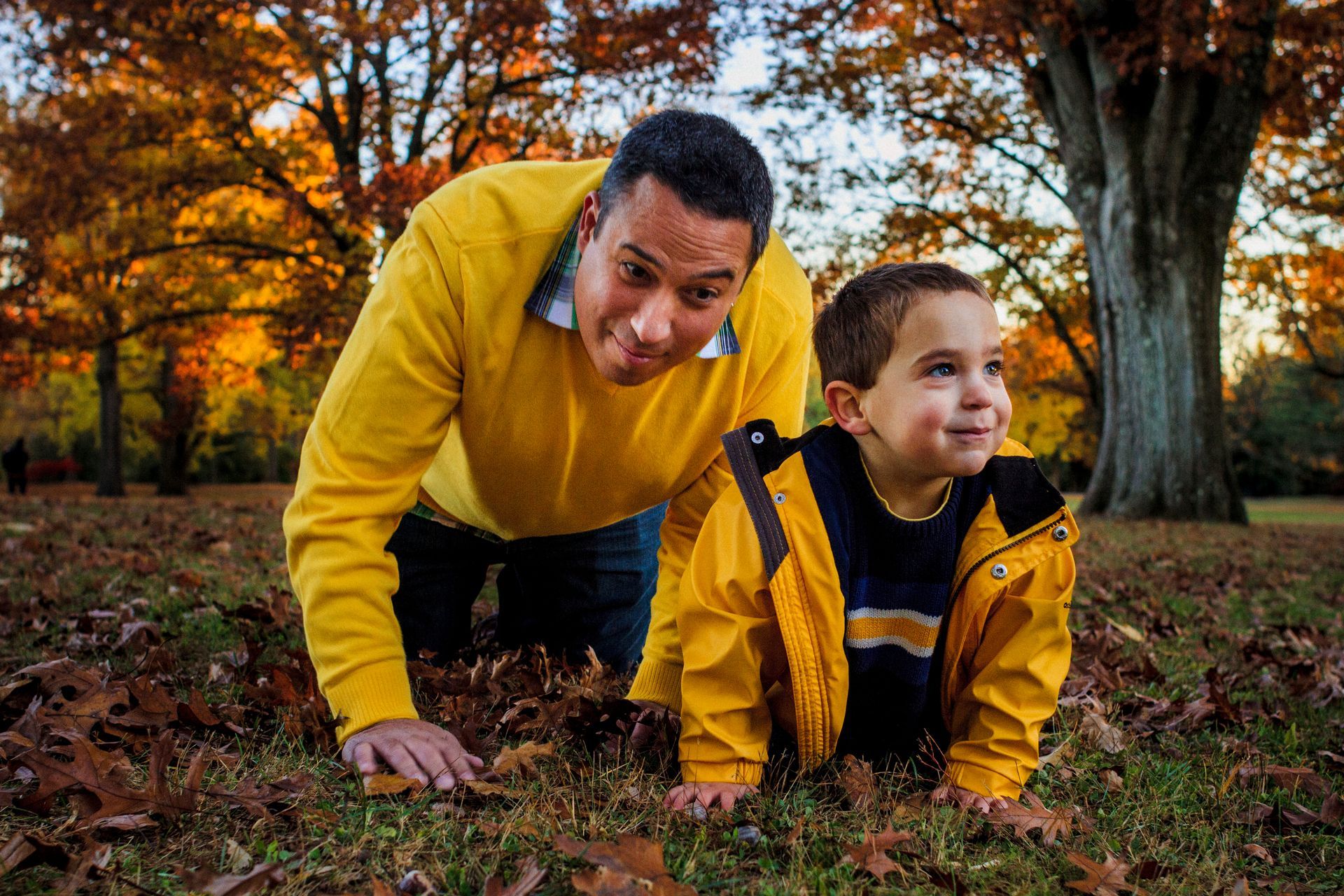 Father and son crawling through fallen leaves, both wearing yellow, outdoors in autumn.