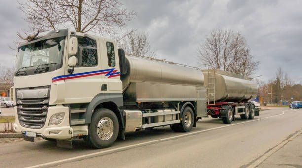 White tanker truck on a road, pulling a trailer. Overcast sky.