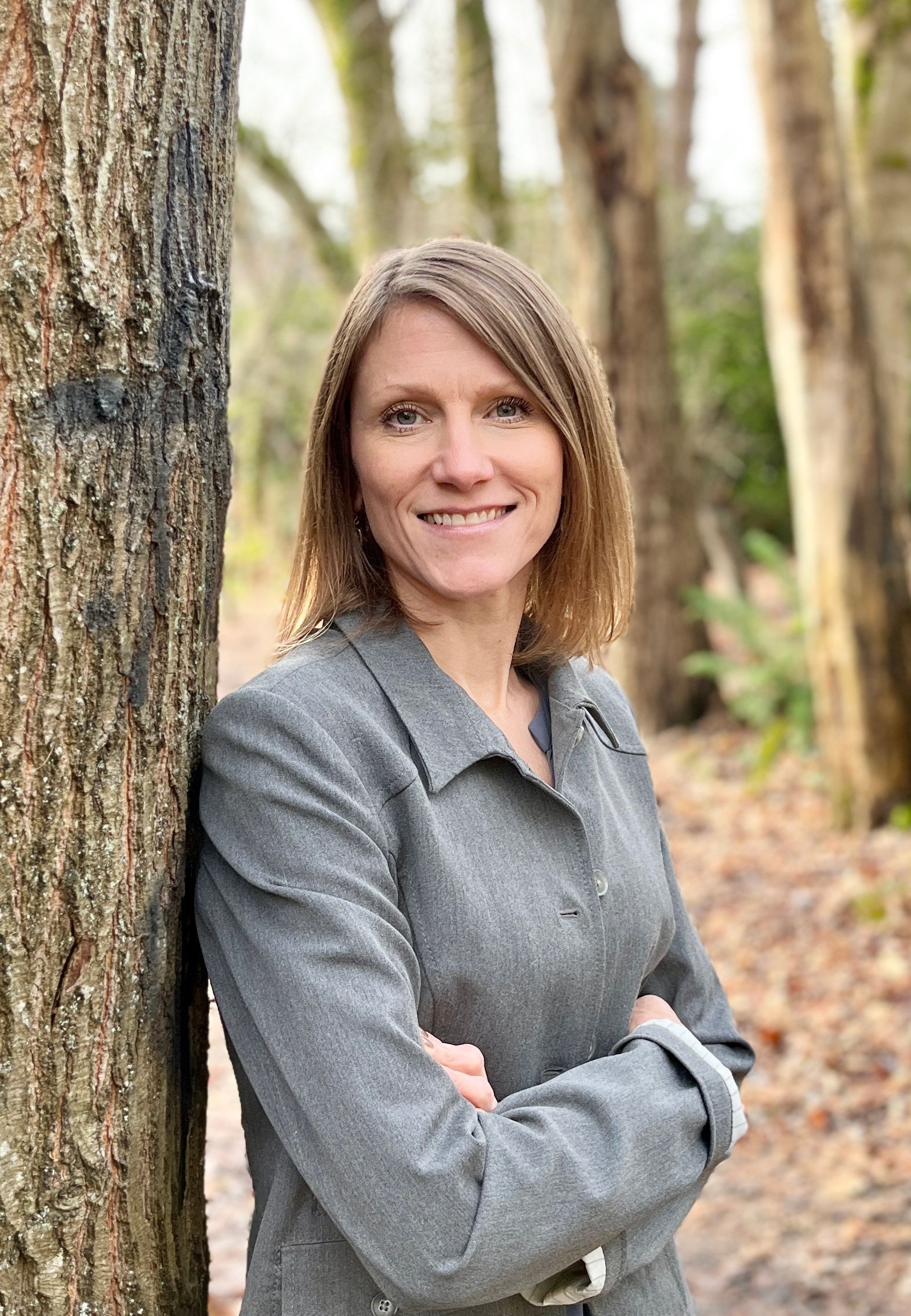 Emily smiles, leaning against a tree in a grey jacket