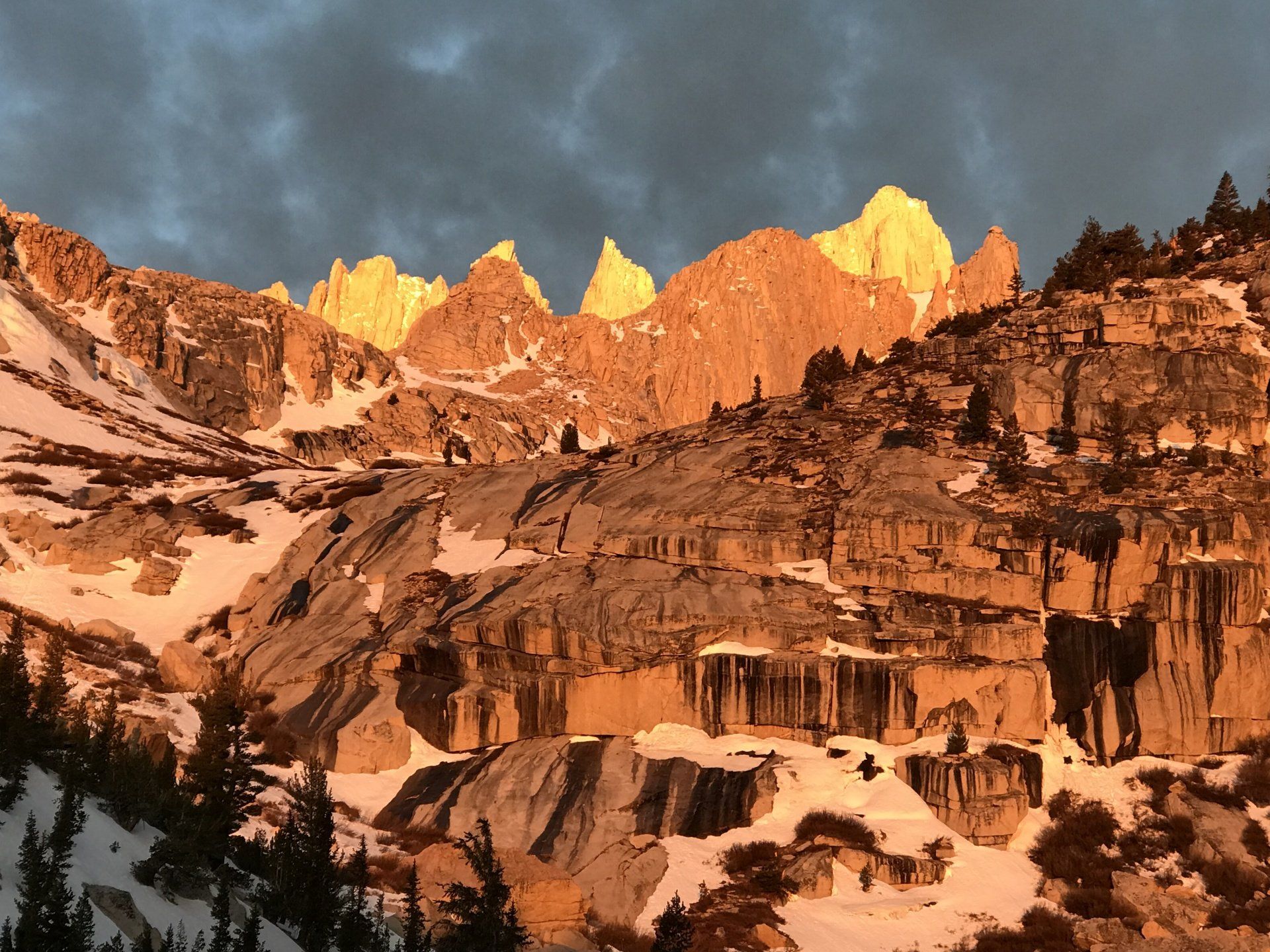 Snowy mountains lit by golden sunset, with dark clouds above.