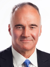 Jacques in dark suit and blue tie, smiling, against white background.