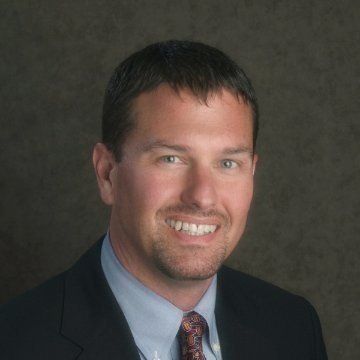 Mike Montgomery in a suit smiles at the camera, against a dark background.