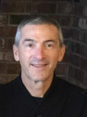 Paul Abdalla with graying hair smiles, posing in front of a brick wall. He wears a black top.