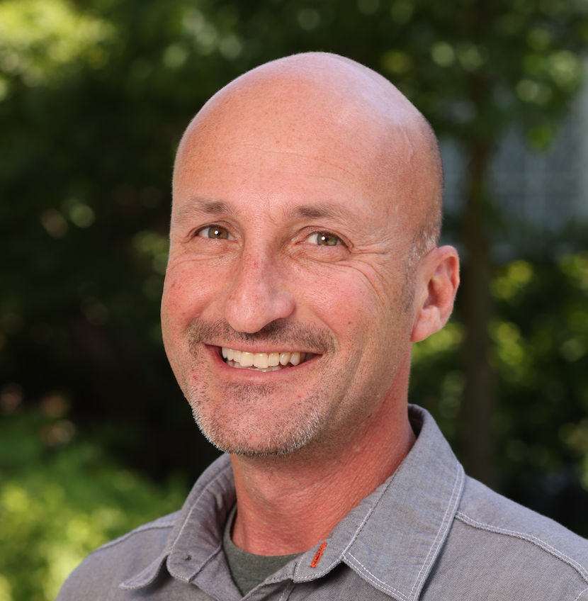 Pete Stamper smiles, wearing a grey collared shirt, in an outdoor setting.