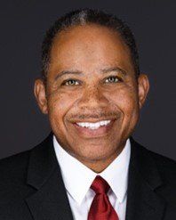 Ron Higgs in a suit smiles at the camera; dark red tie; dark background.