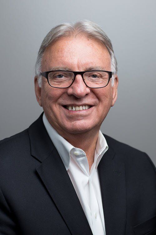 Man in glasses and suit jacket, smiling, neutral gray backdrop.