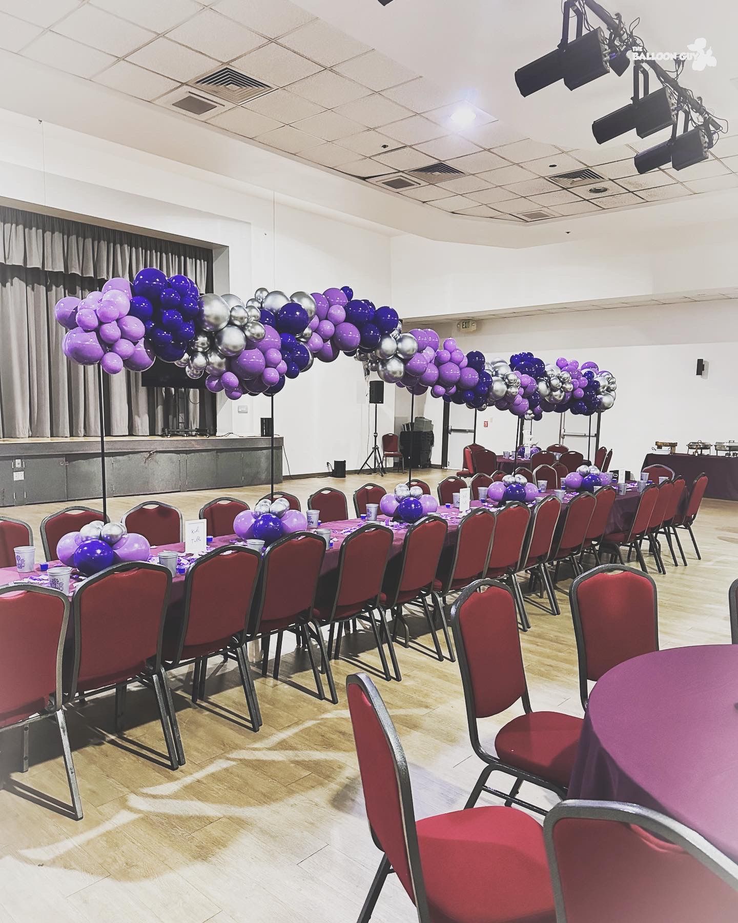 A large room with tables and chairs decorated with purple and silver balloons