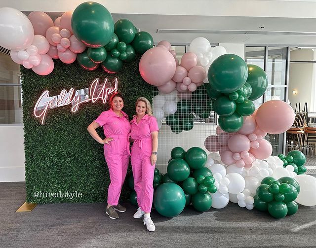 Two women in pink scrubs are standing in front of a wall of balloons.
