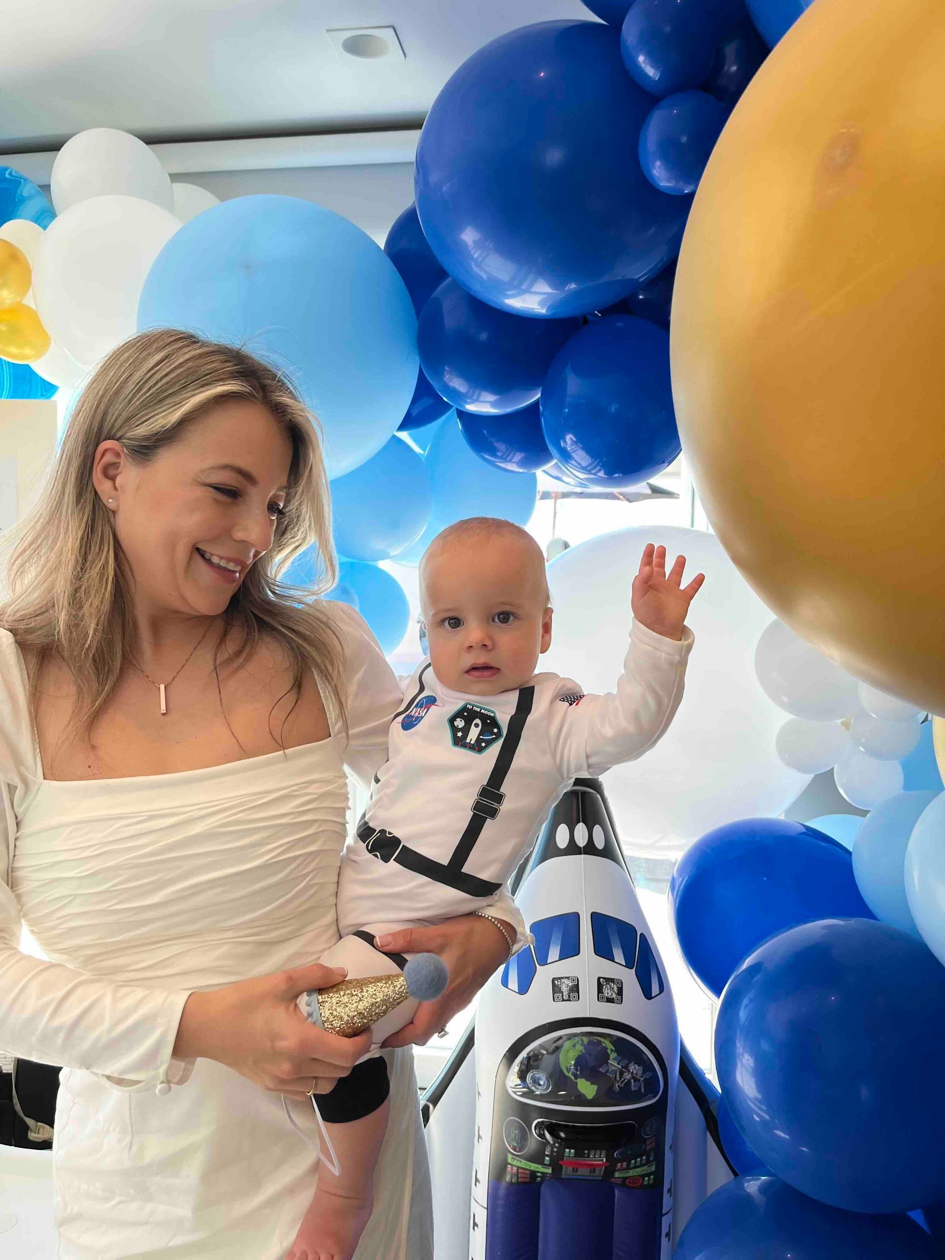 A woman is holding a baby in front of balloons.