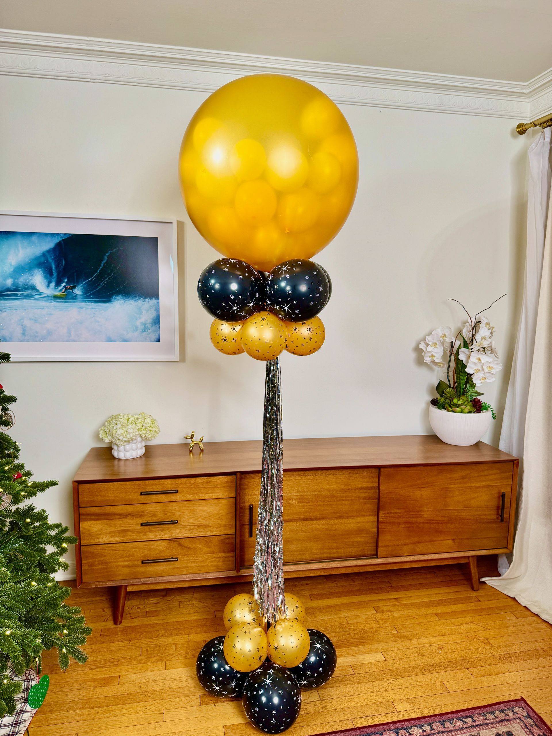 A large gold balloon pop column for New Years Eve is surrounded by black balloons in a living room.