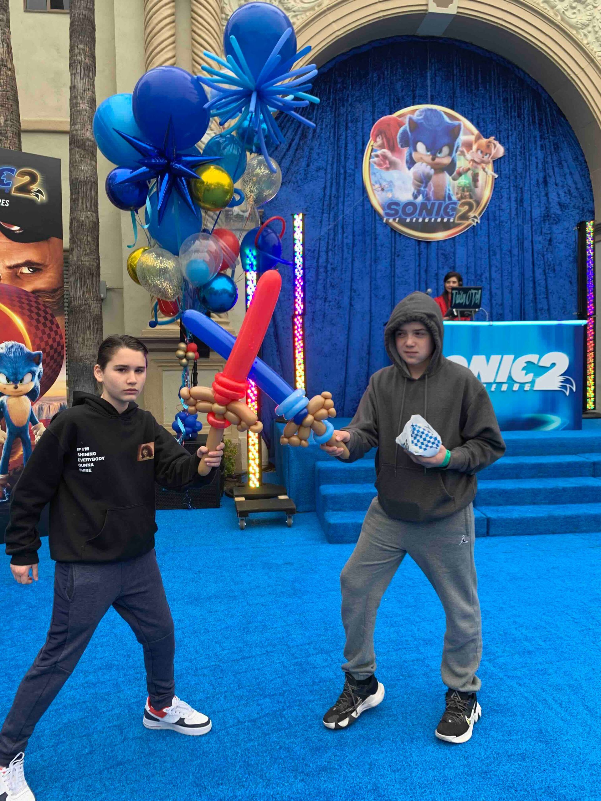 Two boys are standing on a blue carpet with balloons and a sign that says sonic 2