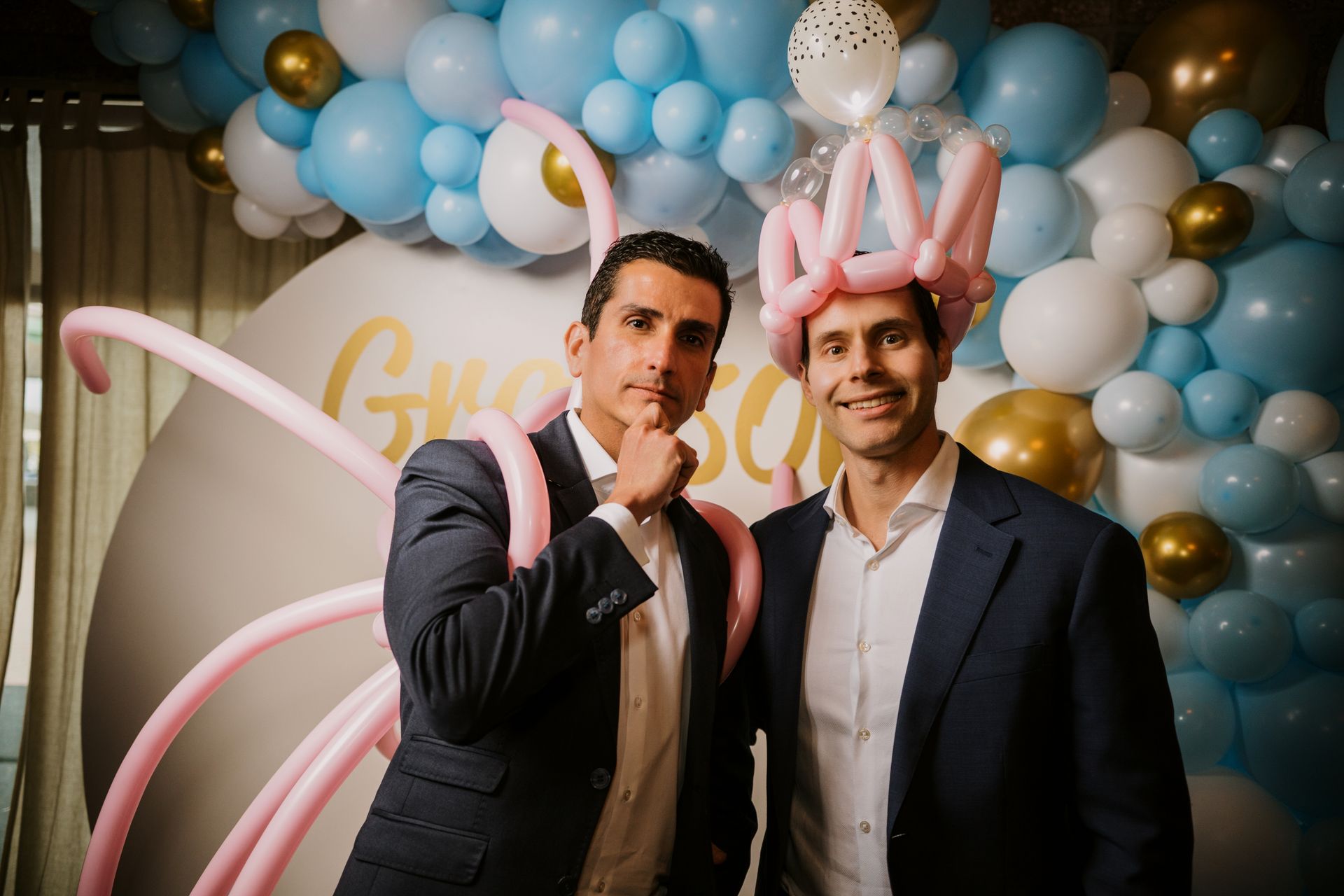 Two men in suits are standing next to each other in front of a wall of balloons.