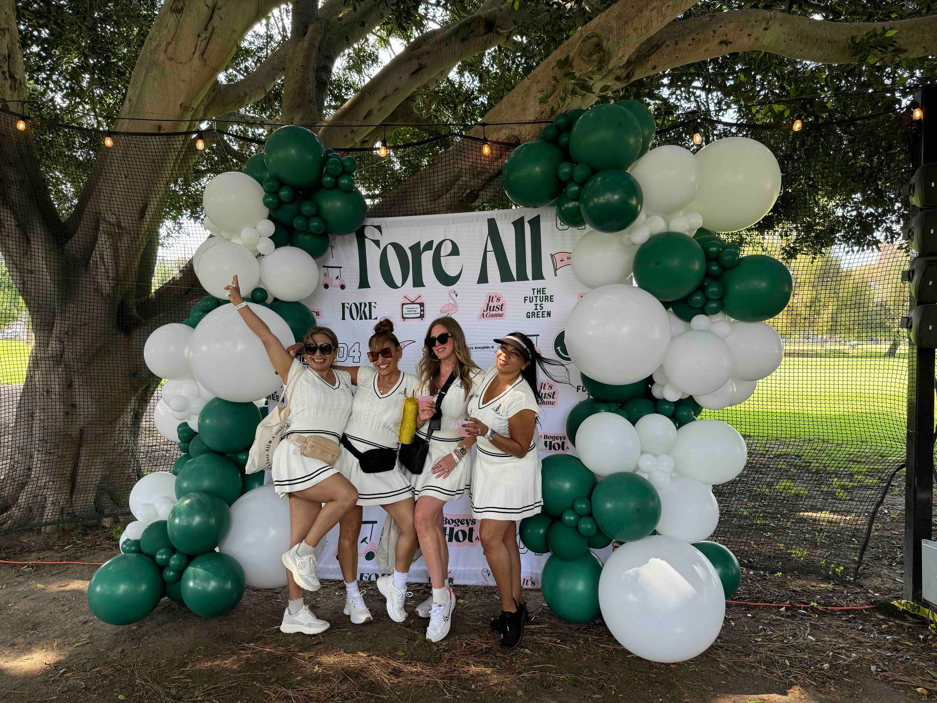 A group of women standing in front of a sign that says `` fore all '' surrounded by green and white balloons.