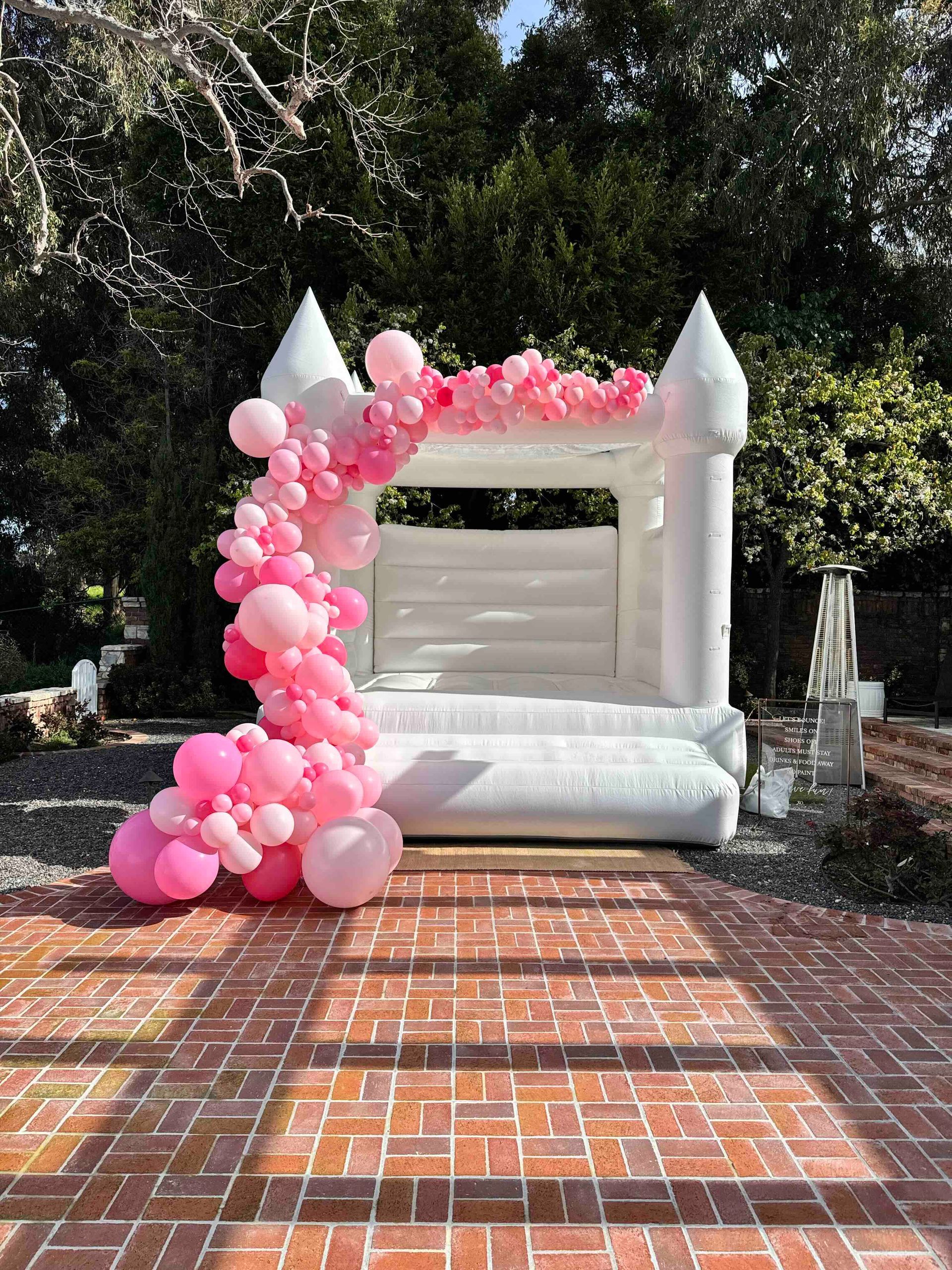 A white bouncy castle surrounded by pink balloons on a brick patio.