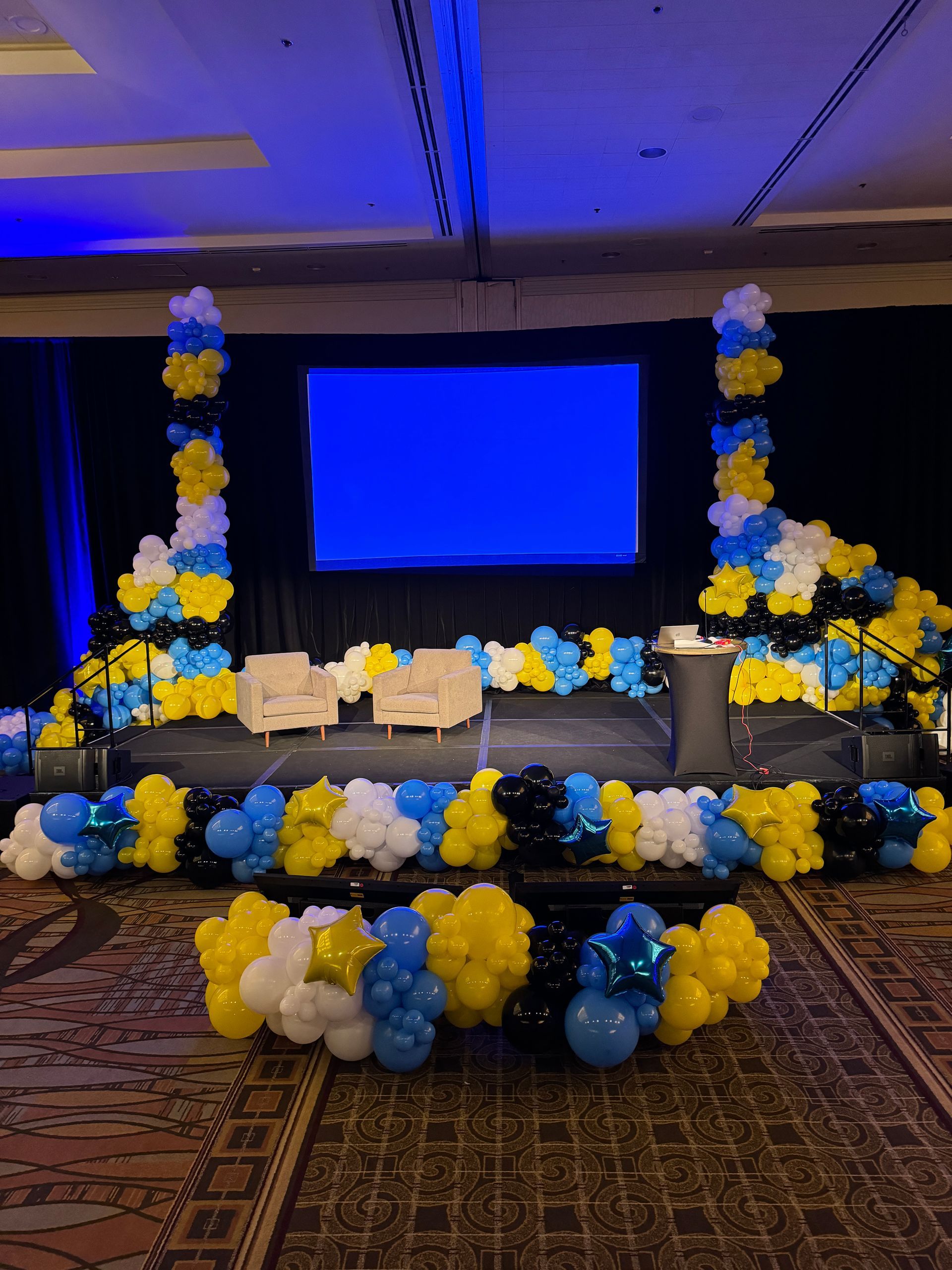 A stage decorated with blue and yellow balloons and a large screen.
