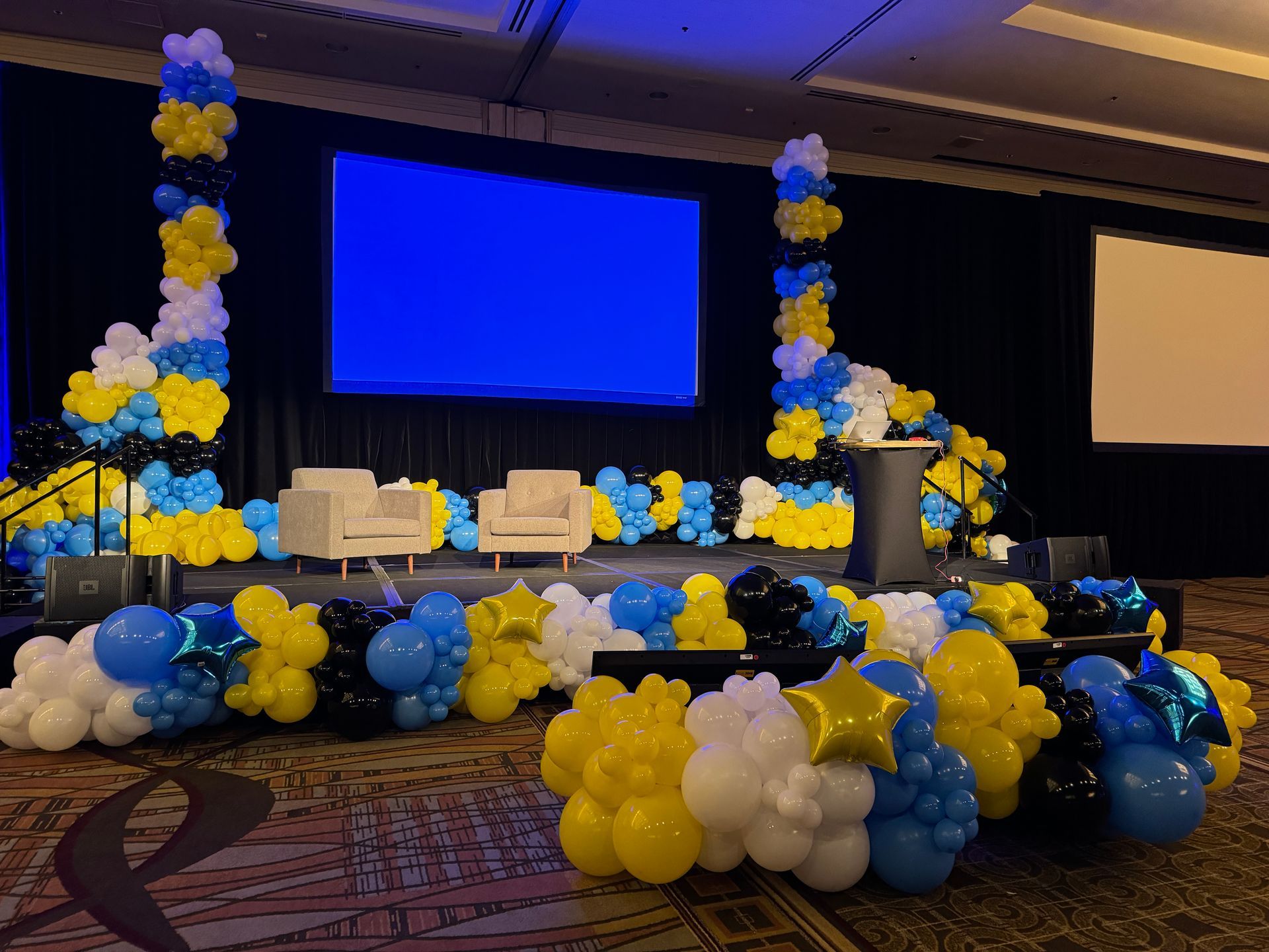 A stage decorated with blue and yellow balloons and a large screen.