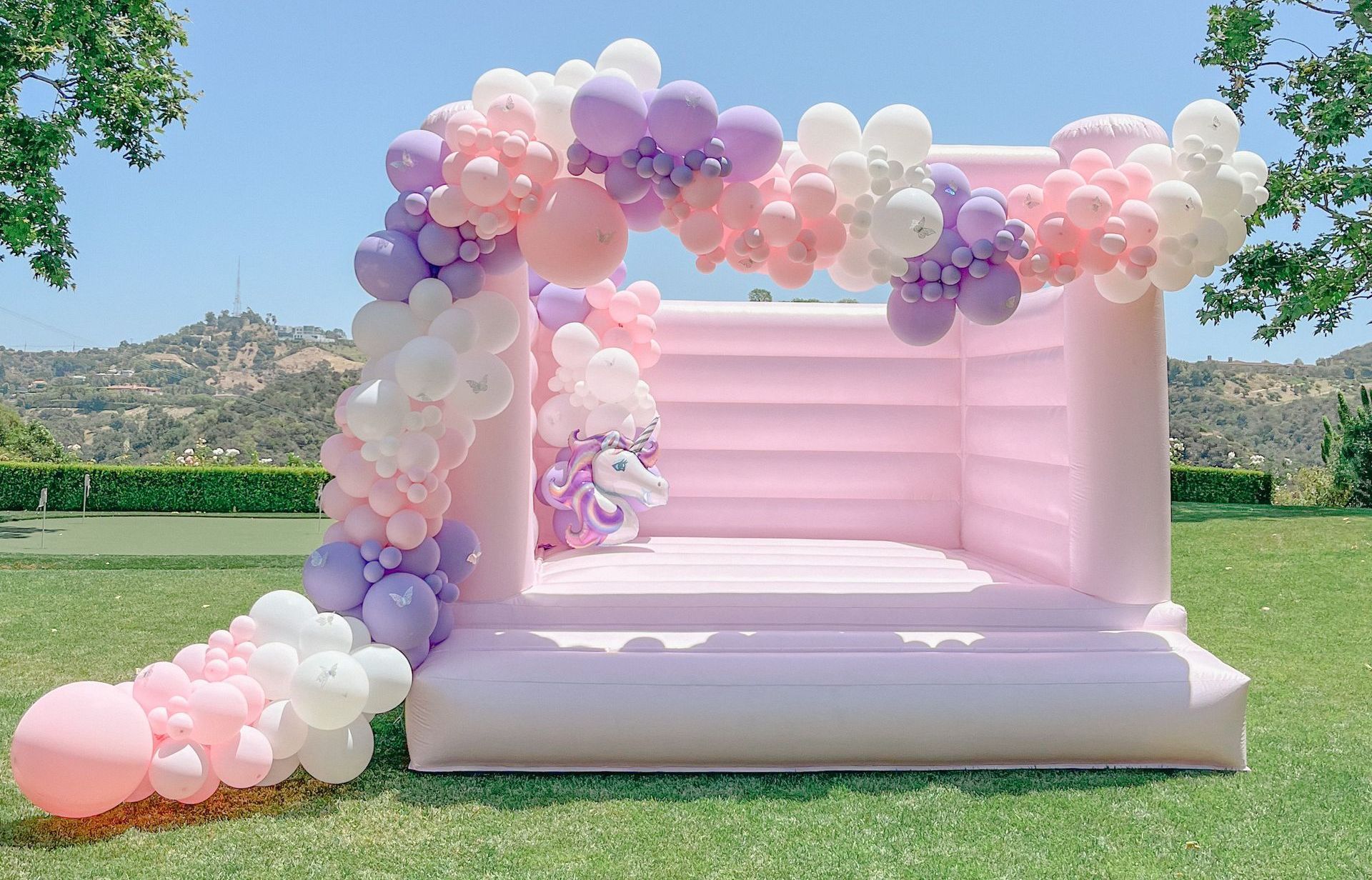 A pink bouncy castle is decorated with pink , white and purple balloons.