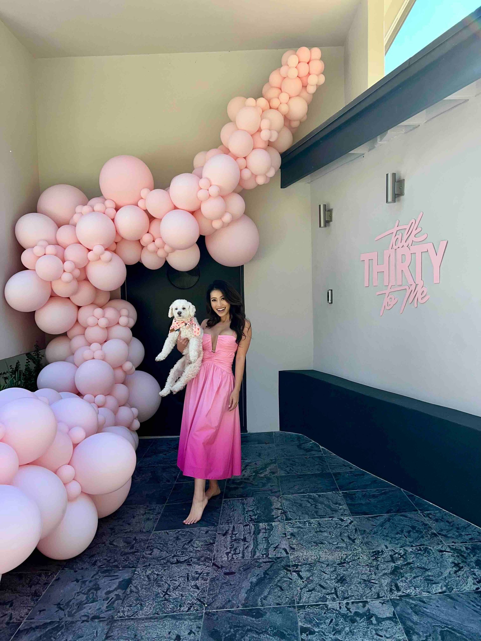 A woman with a dog shows off her dress and pink balloon garland at a 30th birthday party.