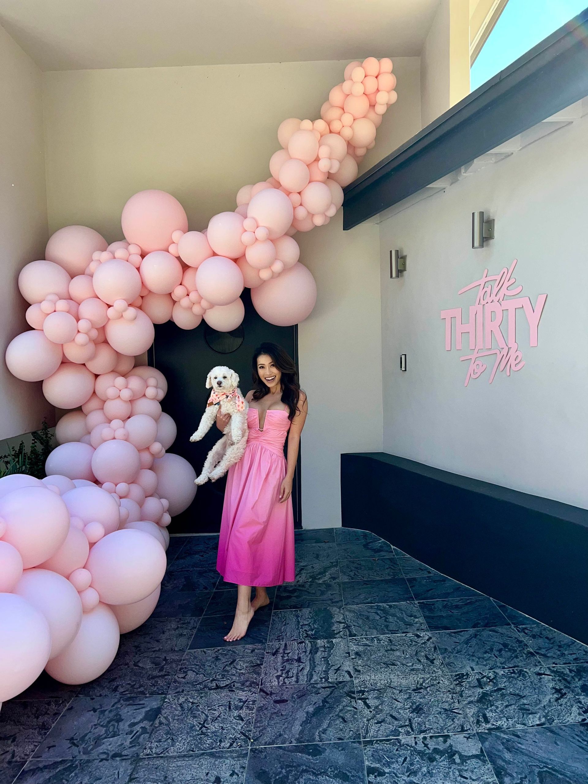 A woman in a pink dress is holding a dog in front of a pink balloon arch.