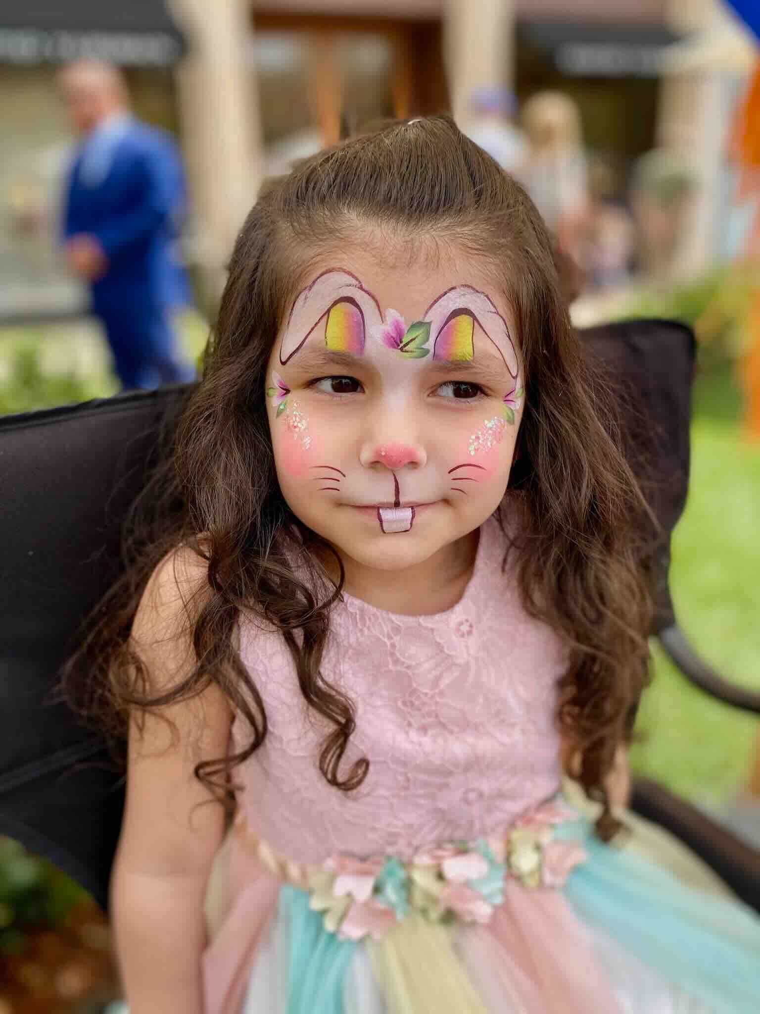 A little girl with her face painted like a bunny is sitting in a chair.