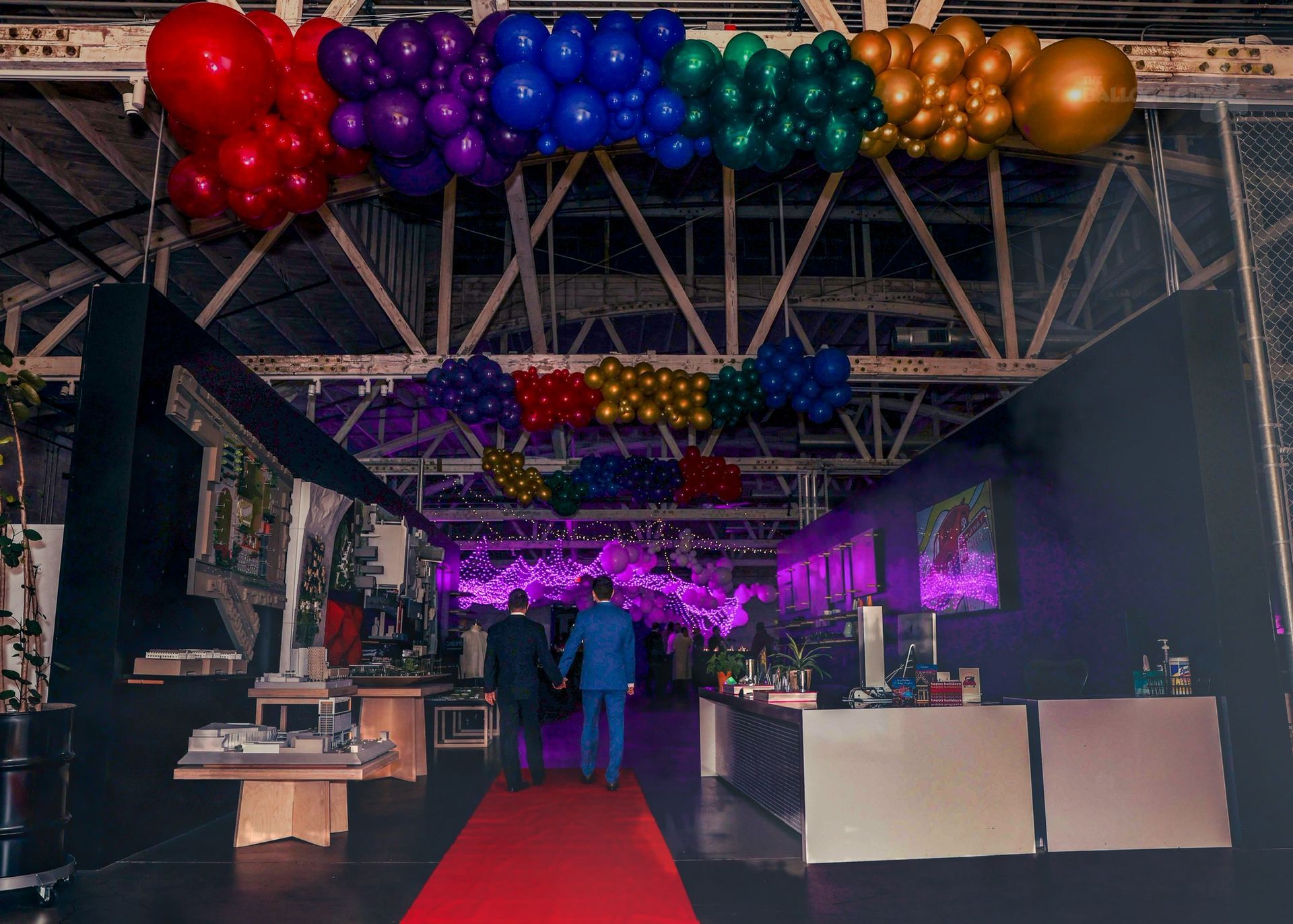A group of people are walking down a red carpet in a room with balloons hanging from the ceiling.