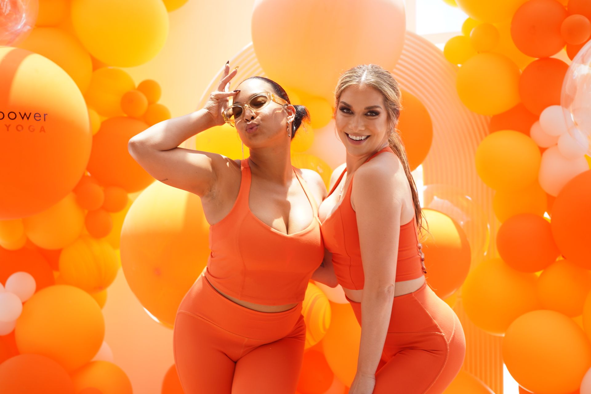 Two women are posing for a picture in front of a wall of orange balloons.