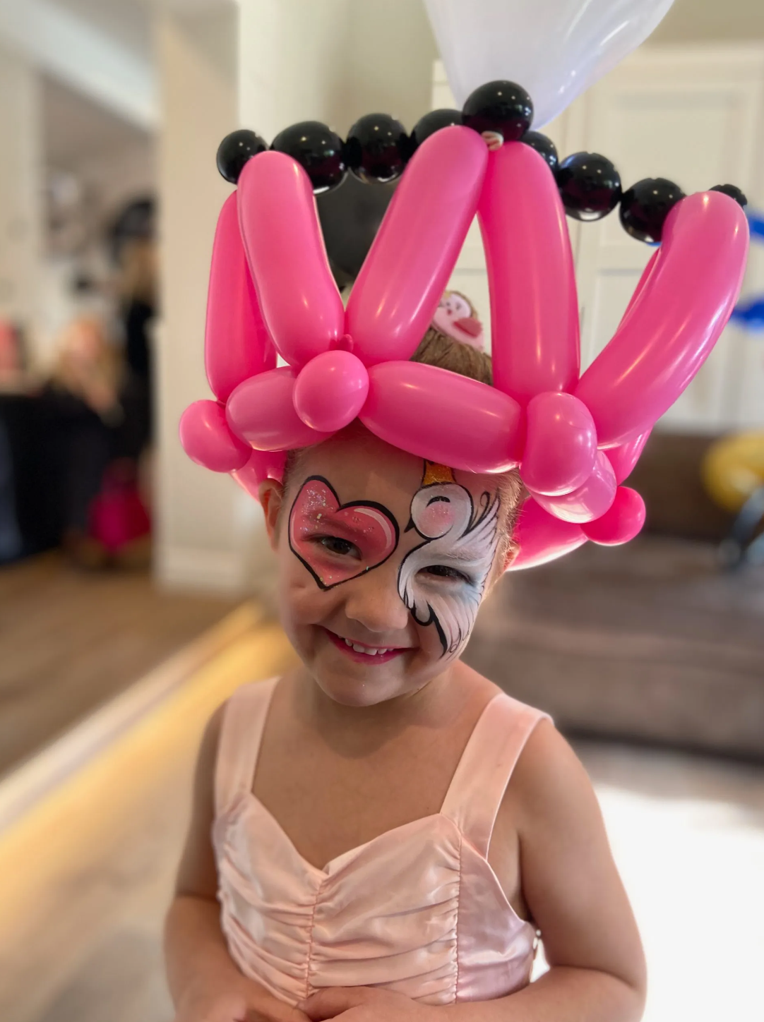 A little girl with her face painted and a hat made of pink balloons.