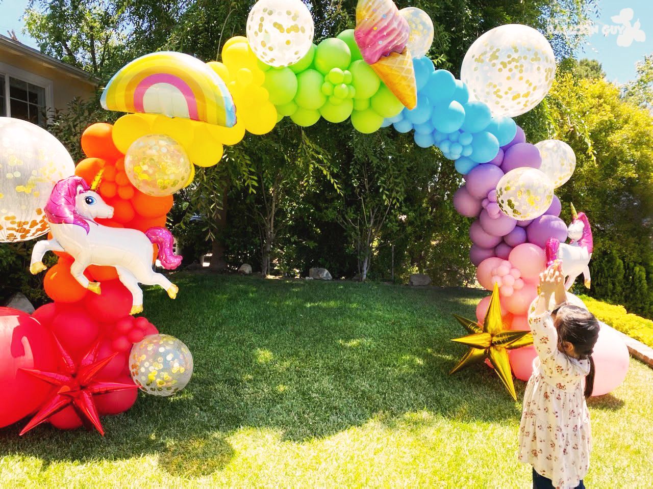 A little girl is standing in front of a rainbow balloon arch.