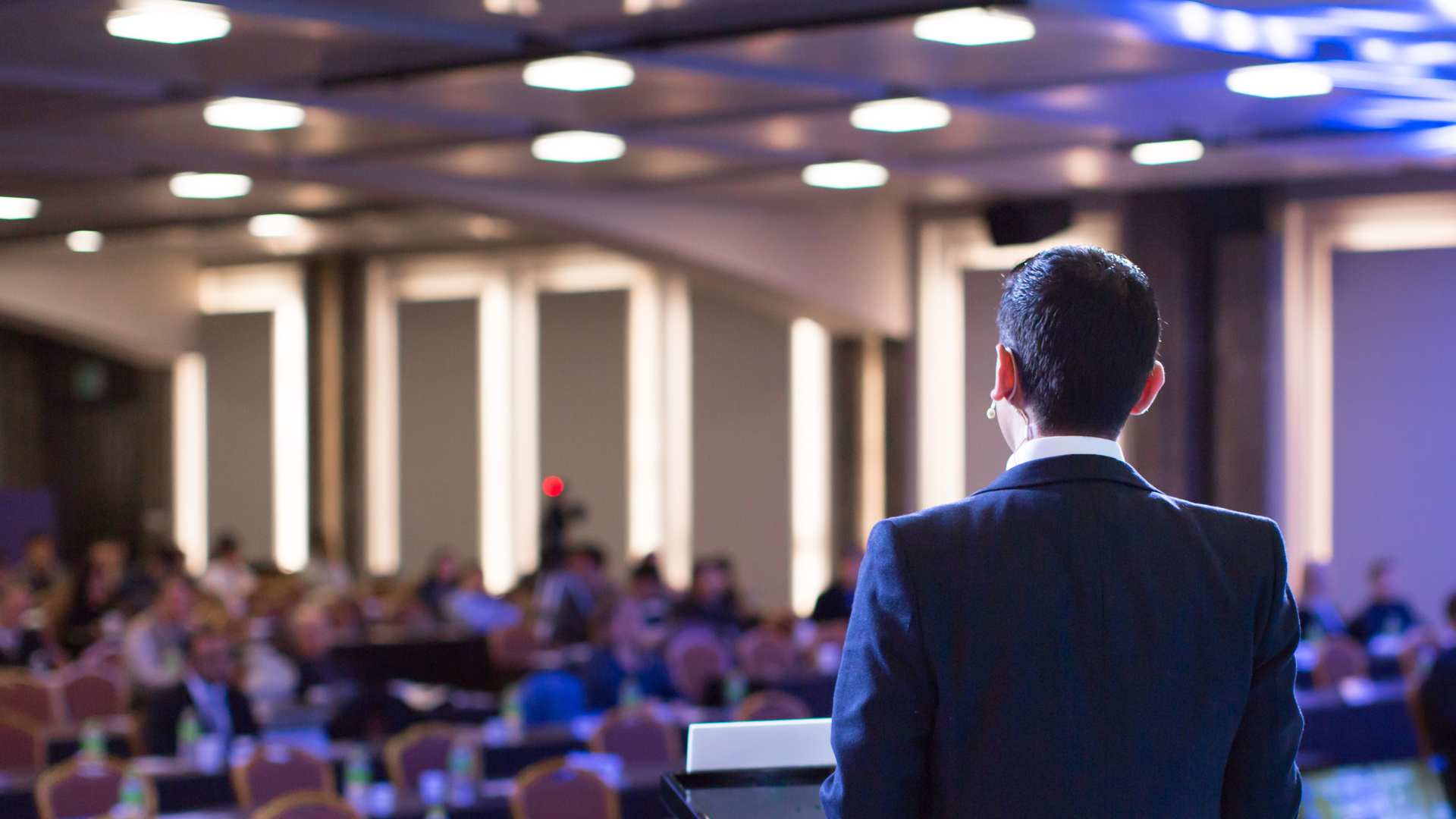 A man in a suit is giving a speech at a podium in front of a crowd.