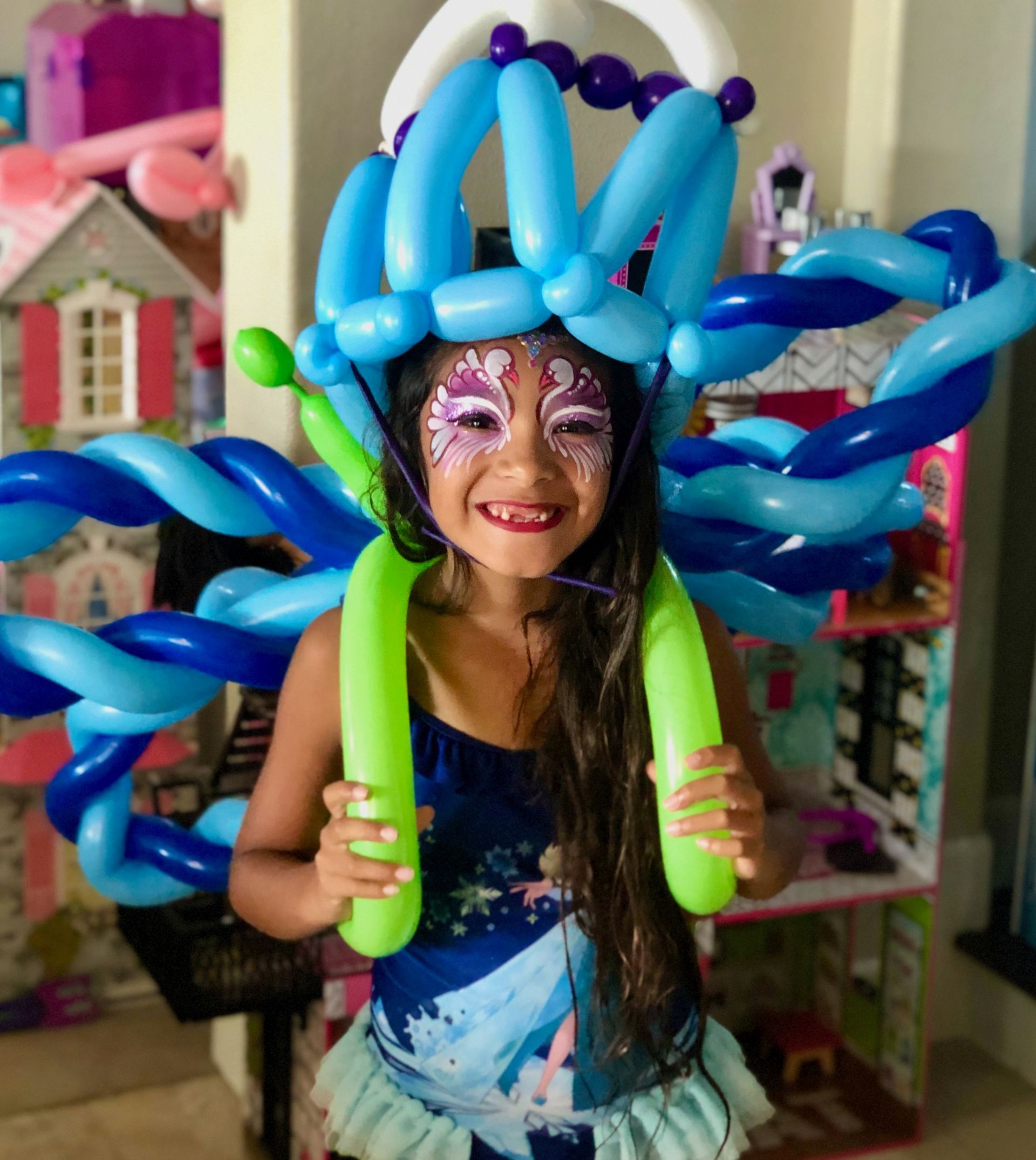 A little girl is wearing a hat and butterfly wings made of blue and green balloons.
