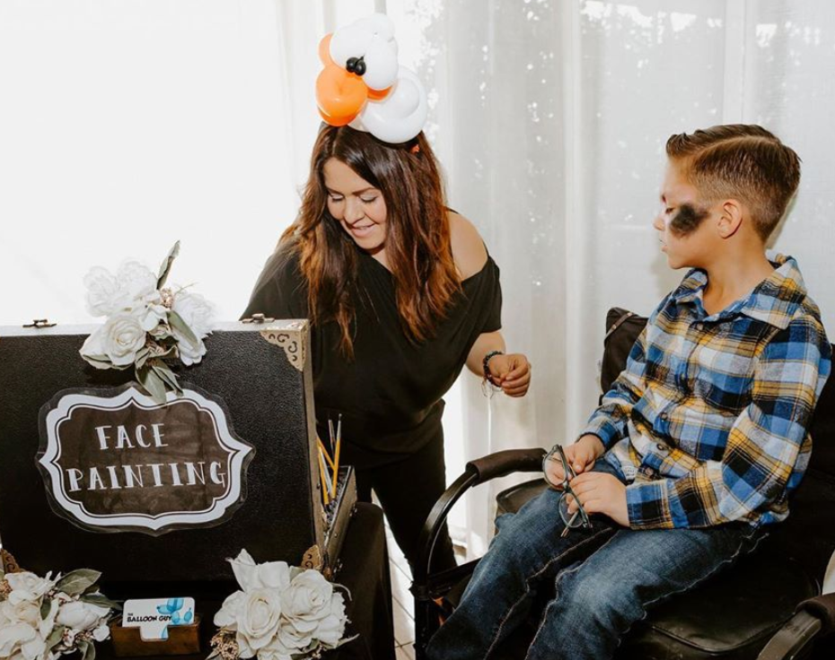 A woman is standing next to a boy in a chair getting his face painted.