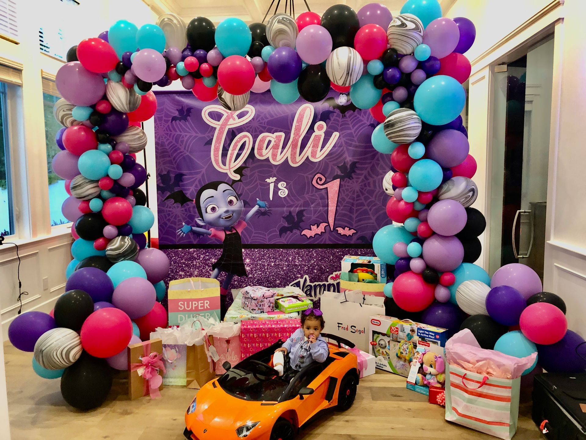 A little girl is sitting in a toy car in front of a balloon arch.