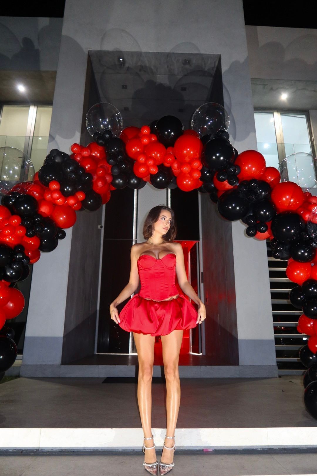 A woman in a red dress is standing in front of a building decorated with red and black balloons.