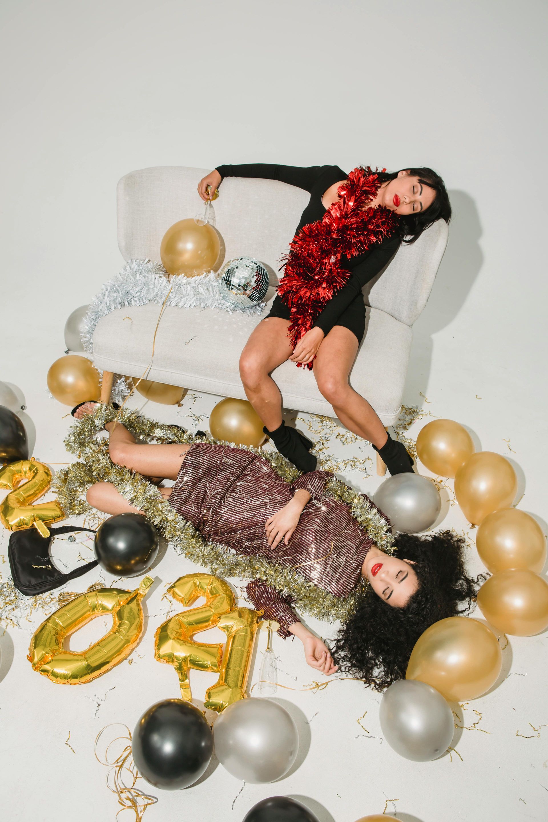 Two women are laying on a couch surrounded by balloons and confetti.