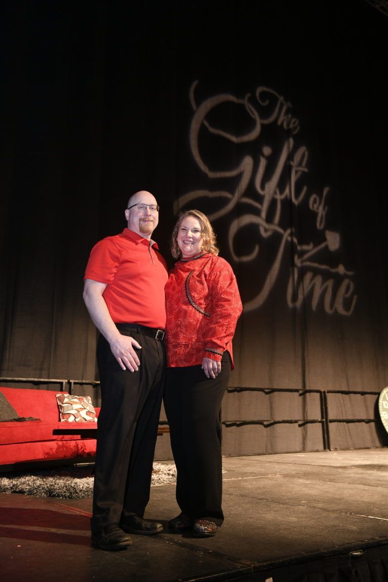 A man and woman are standing on a stage in front of a sign that says the gift of time