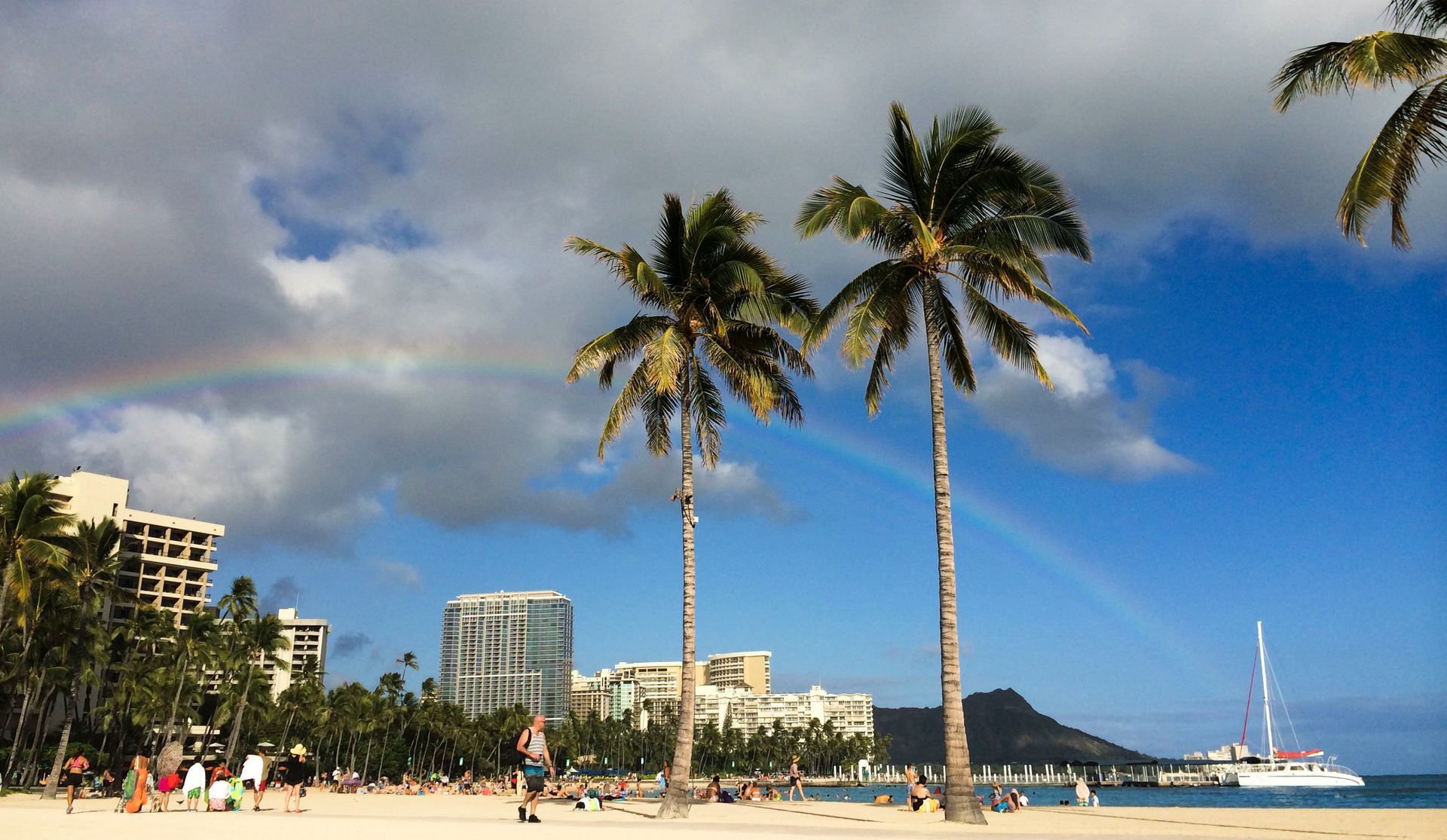 Beach scene with palm trees, rainbow, blue sky, and Diamond Head in the distance.